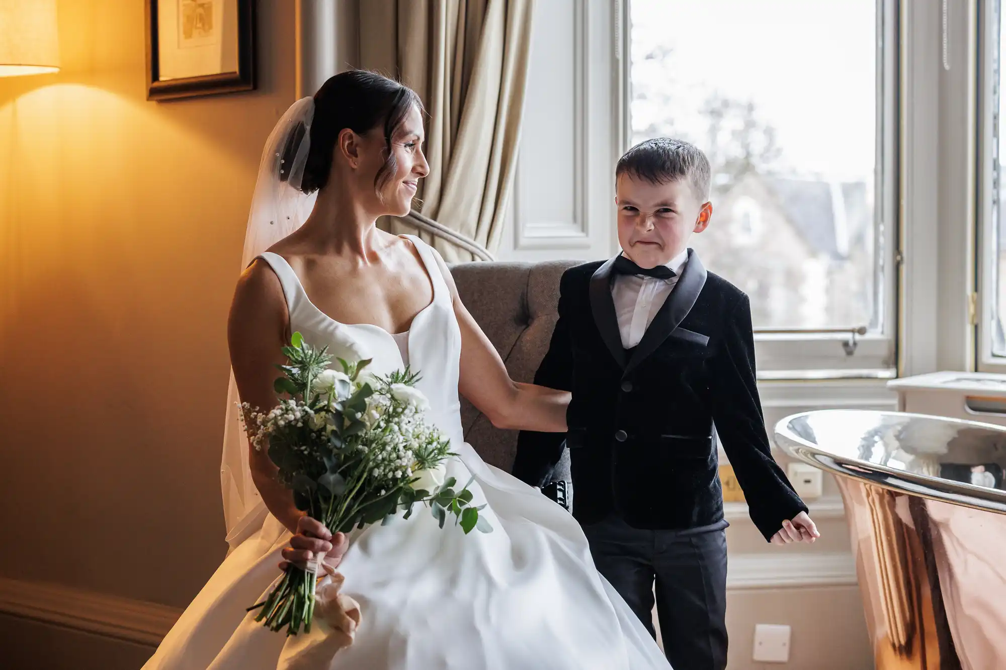 Bride in a white gown holding a bouquet smiles at a young boy in a tuxedo who makes a funny face.