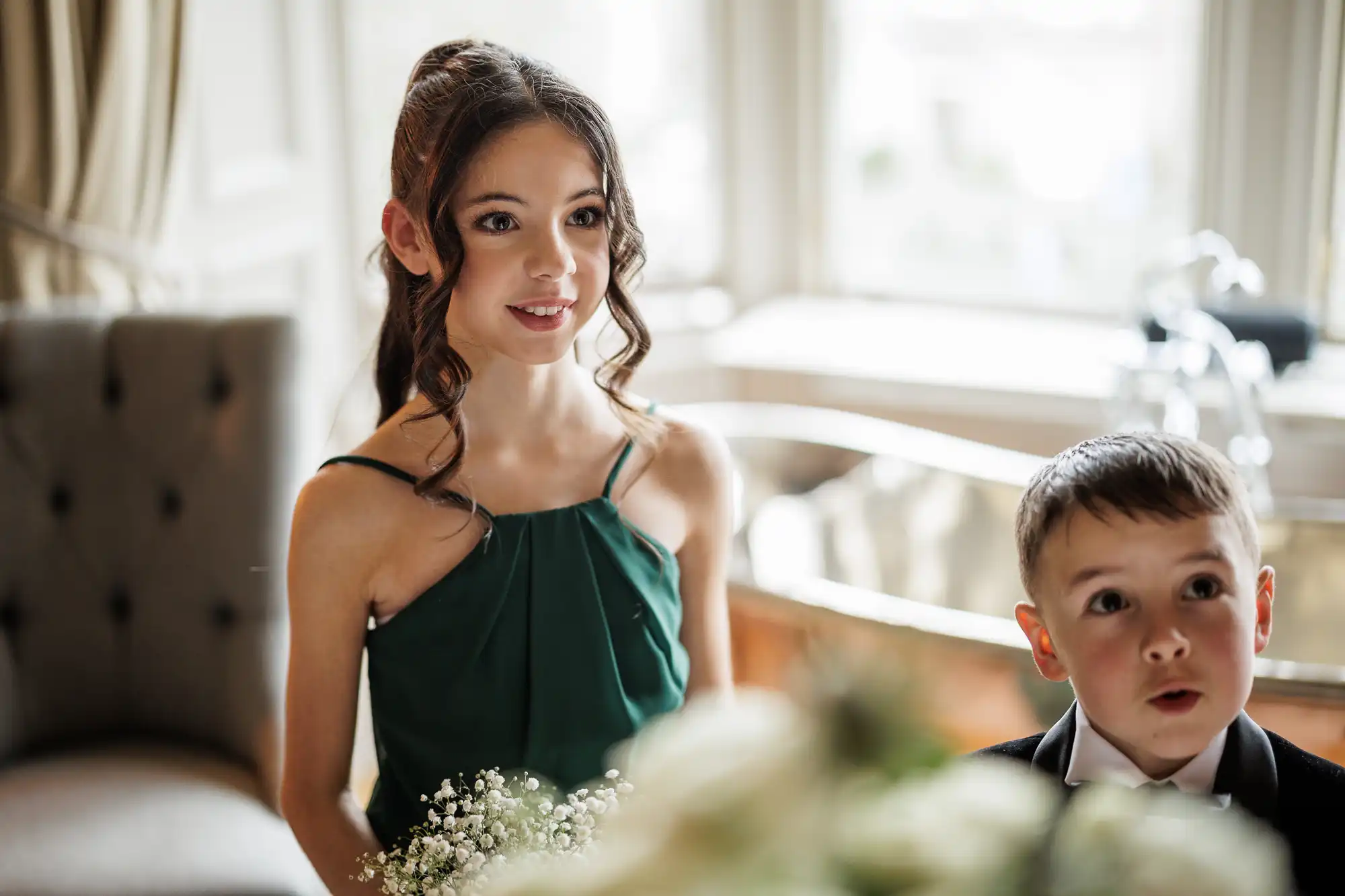 A smiling girl in a green dress holds baby's breath flowers beside a young boy in a black tuxedo with bow tie. Both appear to be in wedding attire, standing indoors near bright windows.