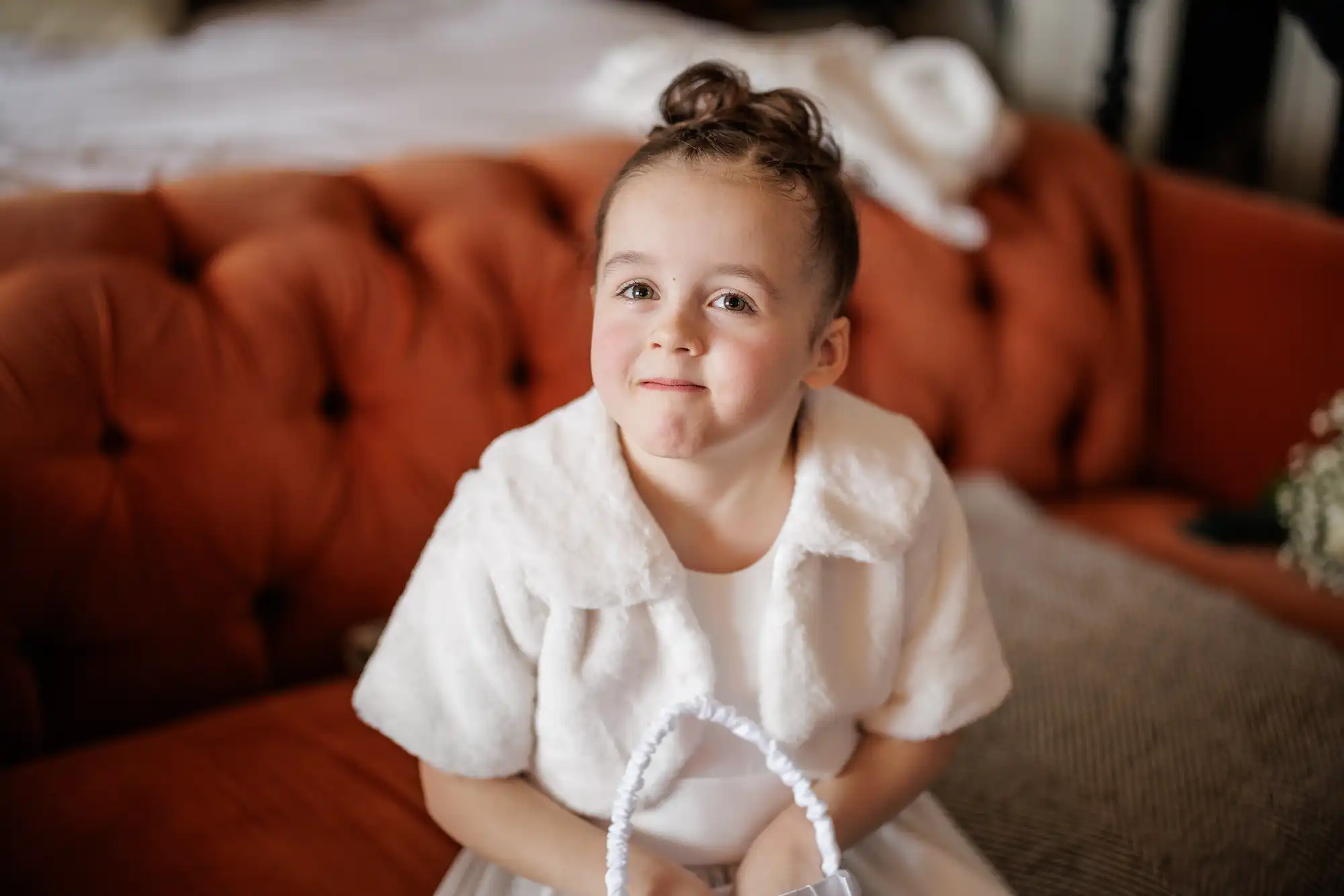 A young girl in a white dress and faux fur shrug sits on an orange sofa, holding a small basket. Her hair is styled in a top knot, and she smiles at the camera.