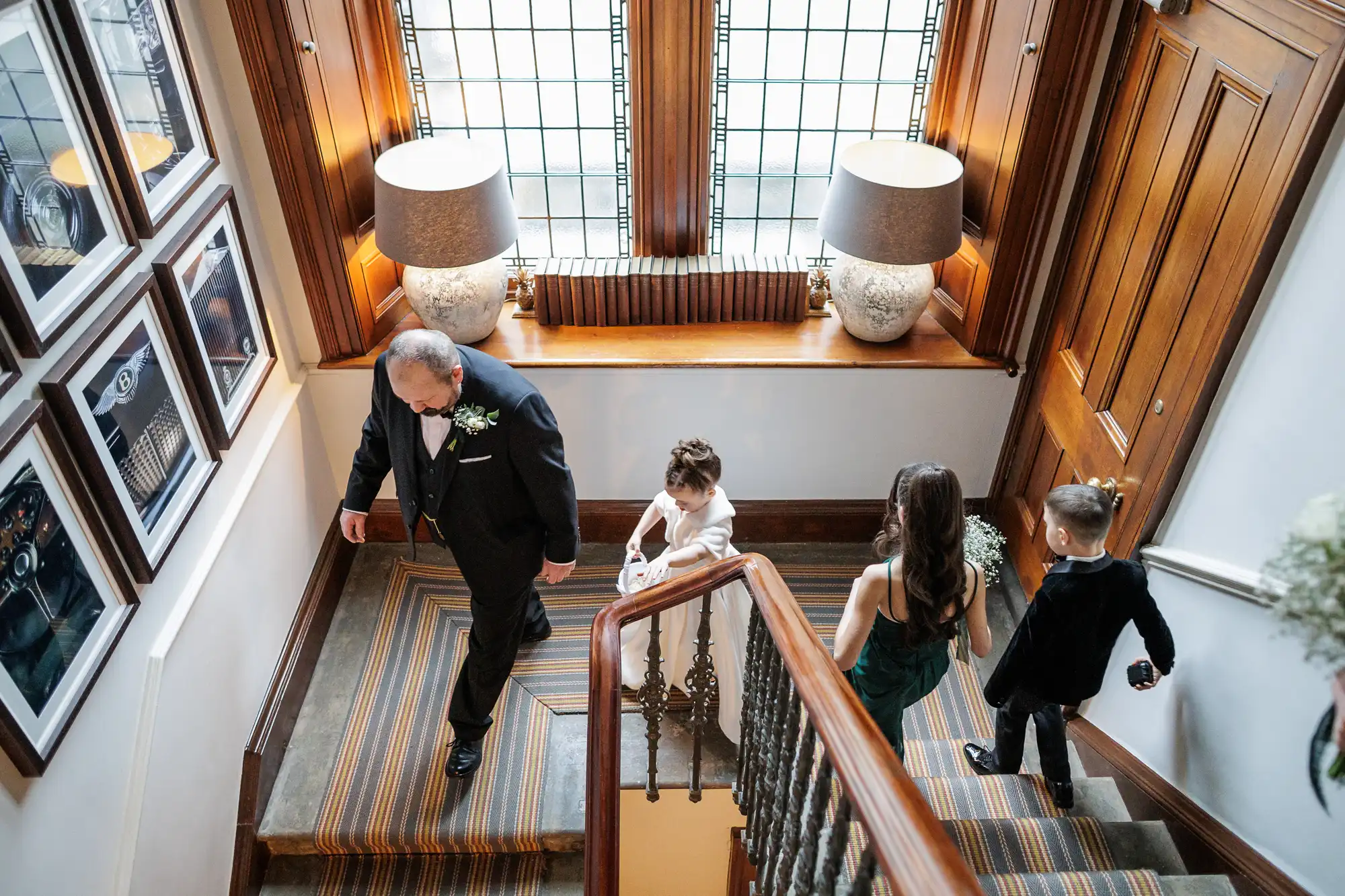 A wedding party walks down a staircase: a groom in a tuxedo, a flower girl in a white dress, a bridesmaid in a green dress, and a ring bearer in a tuxedo.