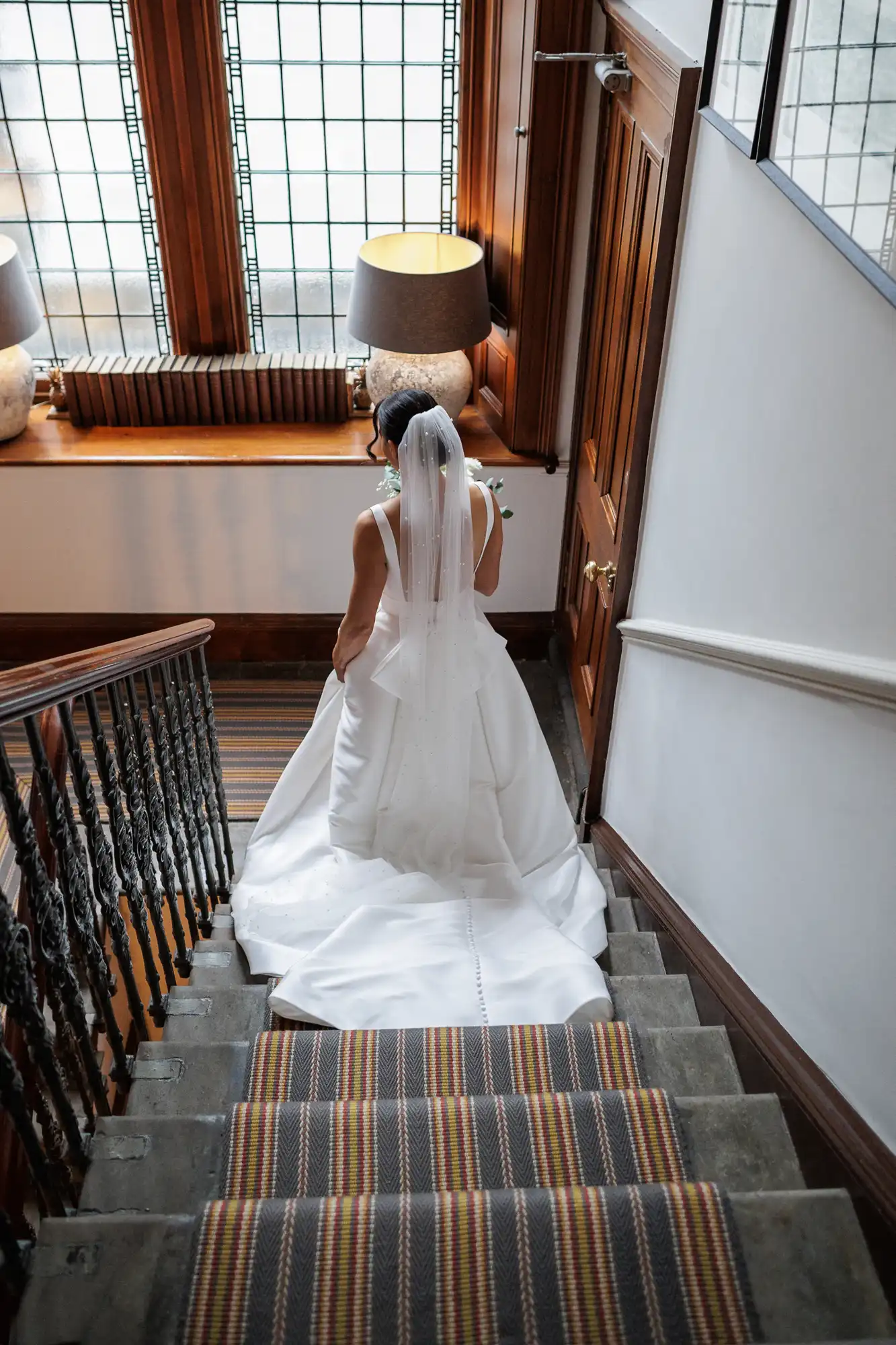 Bride in a flowing white wedding dress and veil walks down a carpeted staircase with ornate railings.