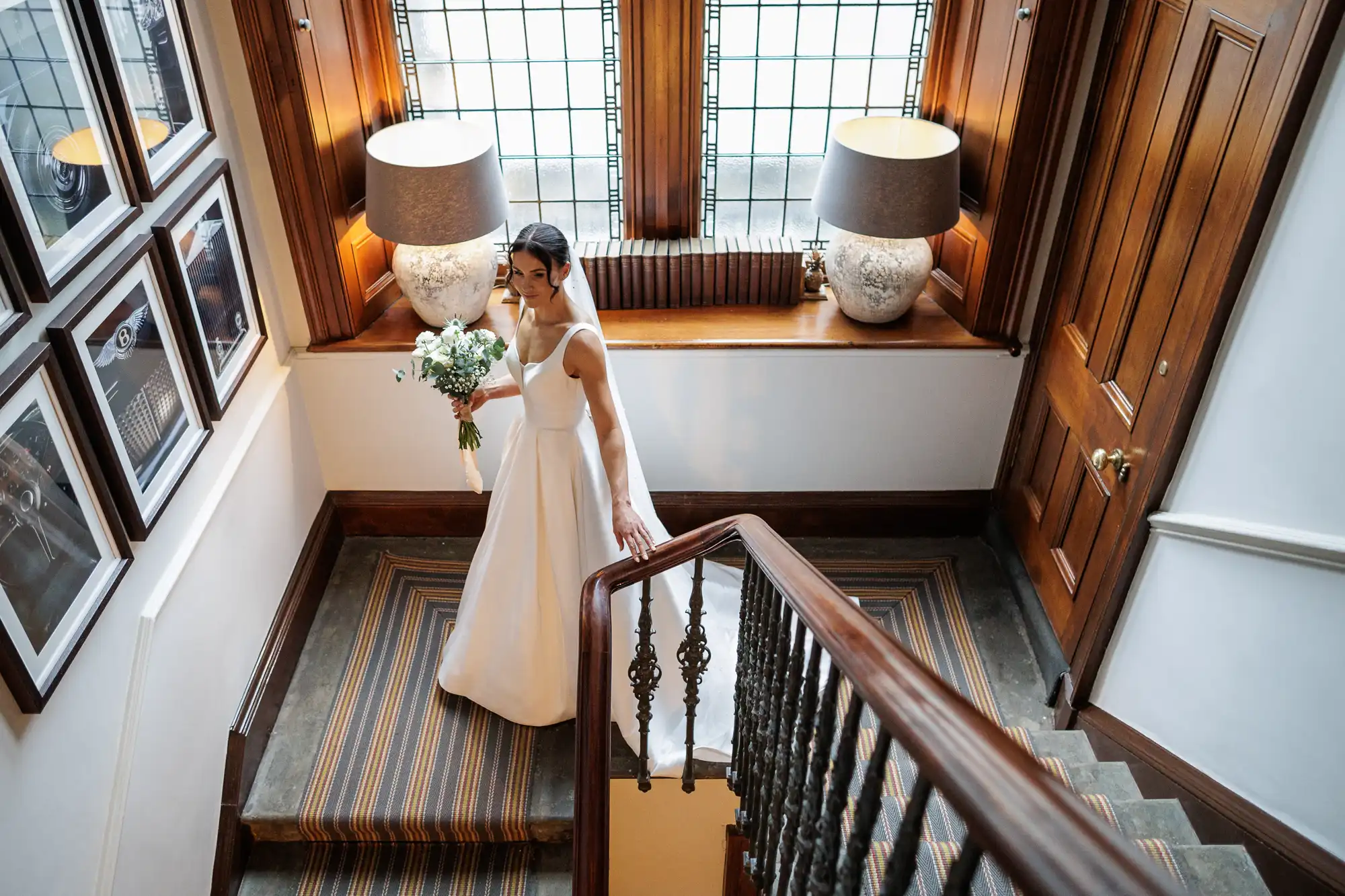 A bride in a white wedding dress, holding a bouquet, walks down a carpeted staircase. She is looking down at her feet. The staircase has a dark wood banister and is lit by two ornate lamps.