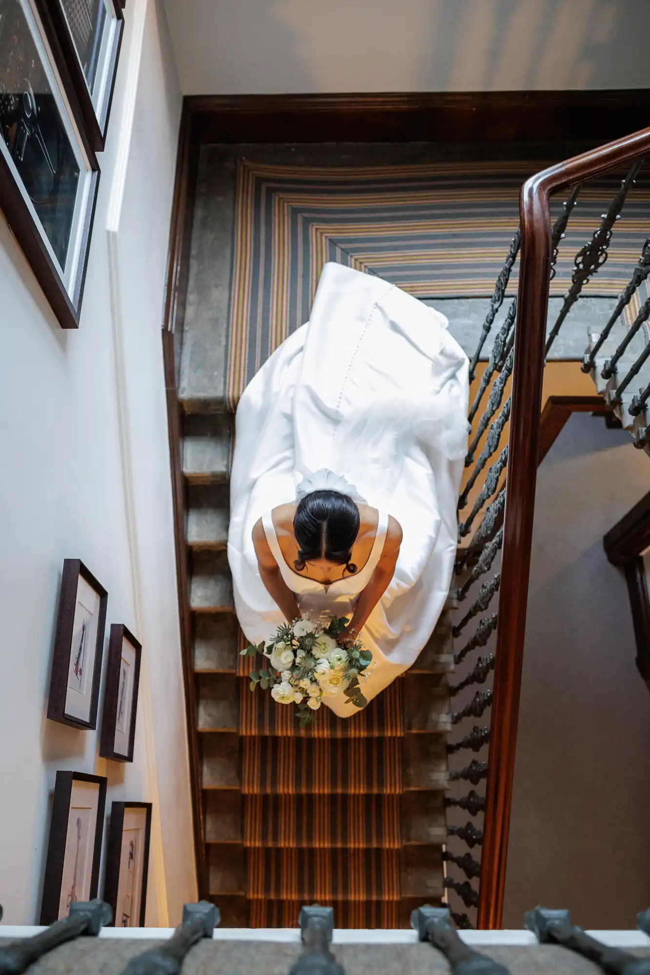A bride in a white wedding dress holding a bouquet of white flowers stands on a staircase. The staircase has a striped runner and a wooden banister.