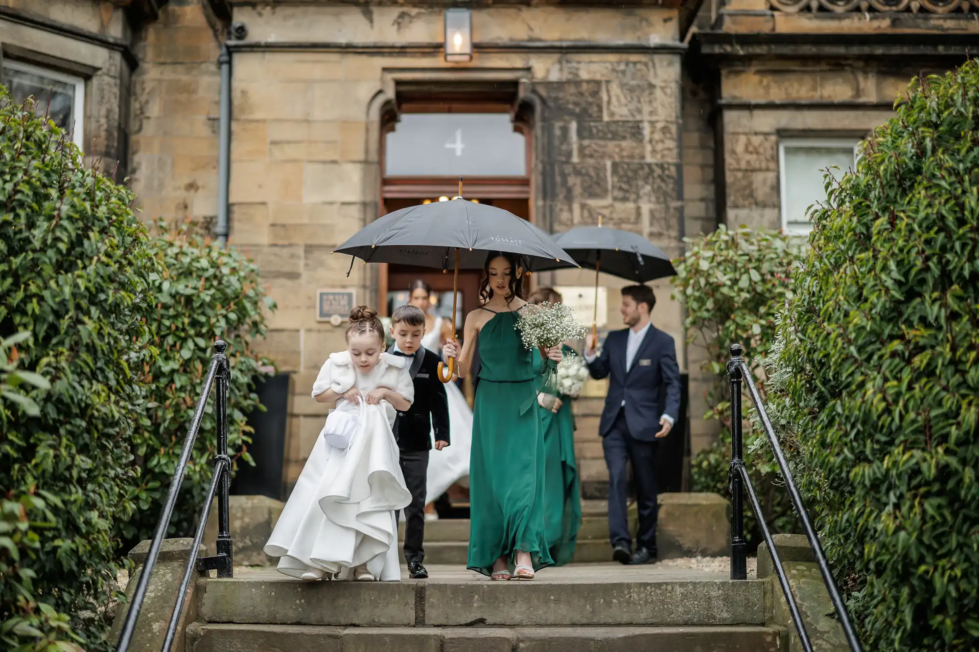 A wedding party walks down stone steps toward the camera under umbrellas on a rainy day. The bride, in a green dress, leads, holding a bouquet. A flower girl in a white dress and a page boy in a tuxedo follow closely behind.