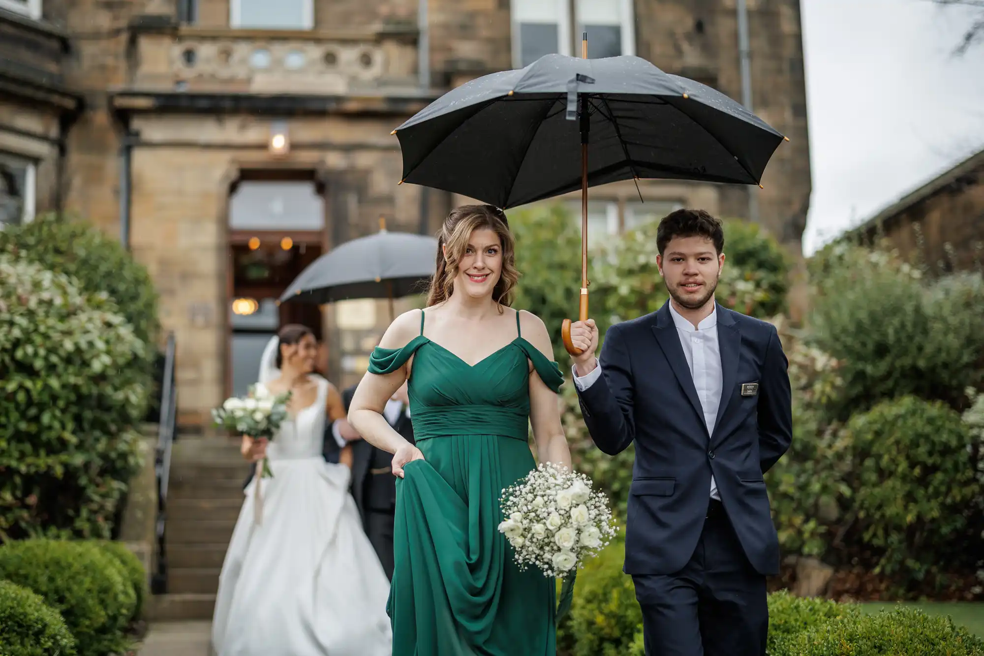 A bridesmaid in a green dress holds a bouquet under a black umbrella as she walks with a groom in a navy suit. A bride in a white gown is in the background.