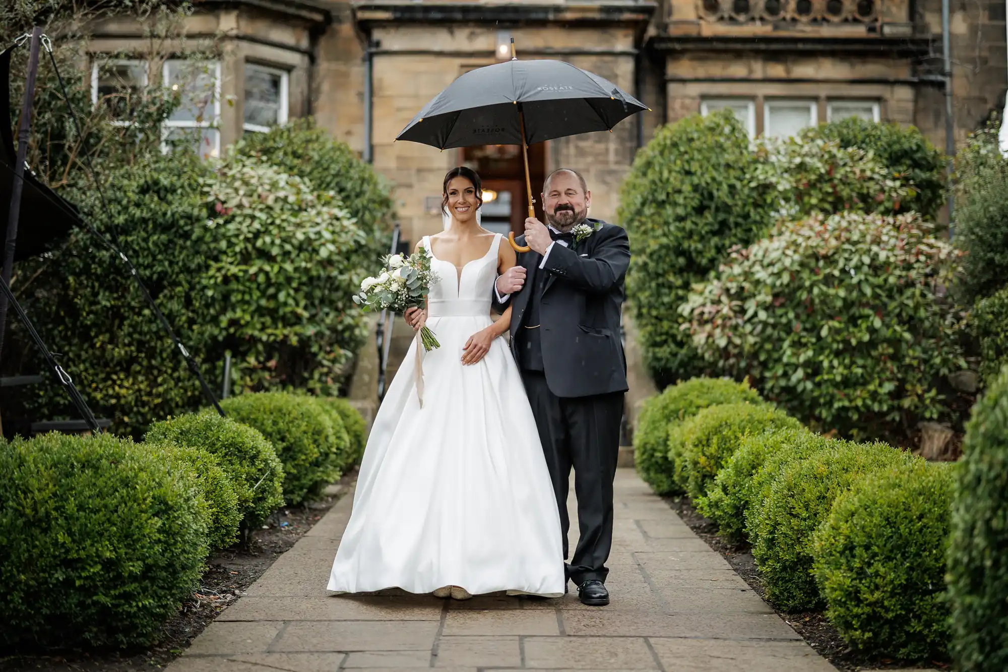 A smiling bride in a white gown and a groom in a black suit share an umbrella on a paved path lined with manicured bushes.