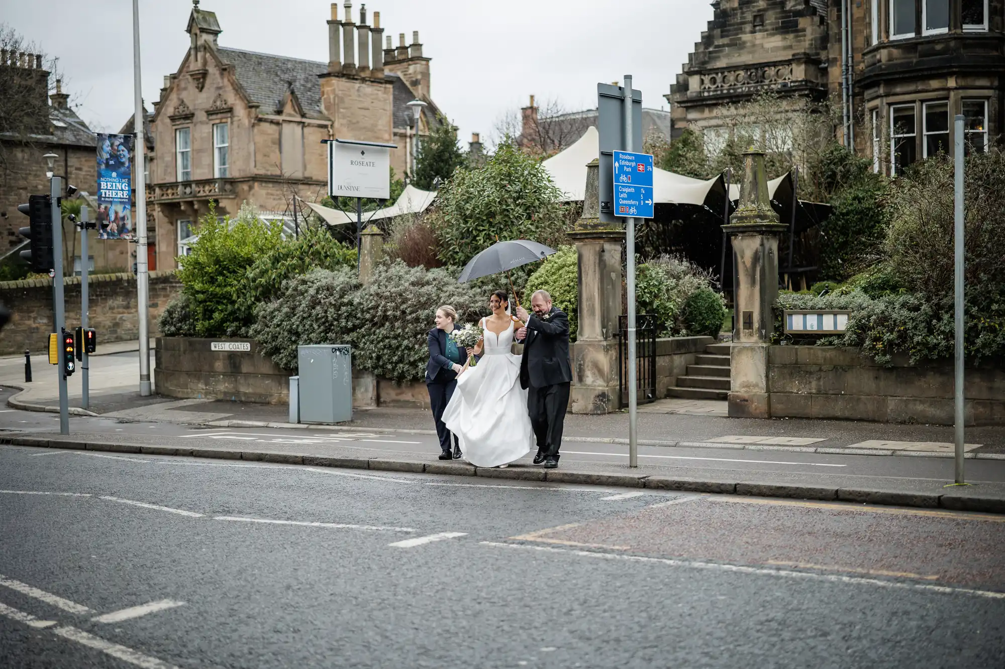 A bride in a white dress walks with an umbrella under her arm, escorted by two individuals in suits, on a cloudy day.