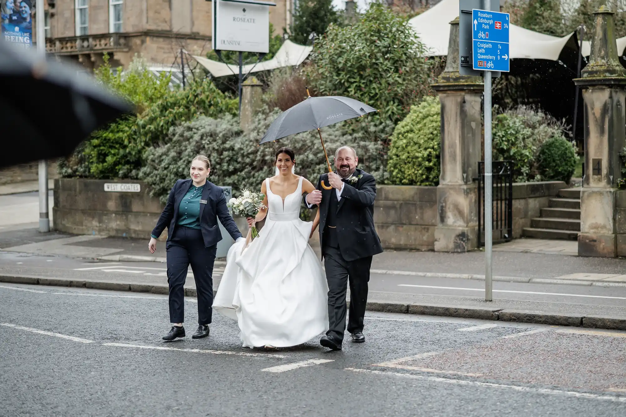 A bride in a white gown and groom in a suit walk under a black umbrella on a rainy day, with a woman in a dark suit by their side.