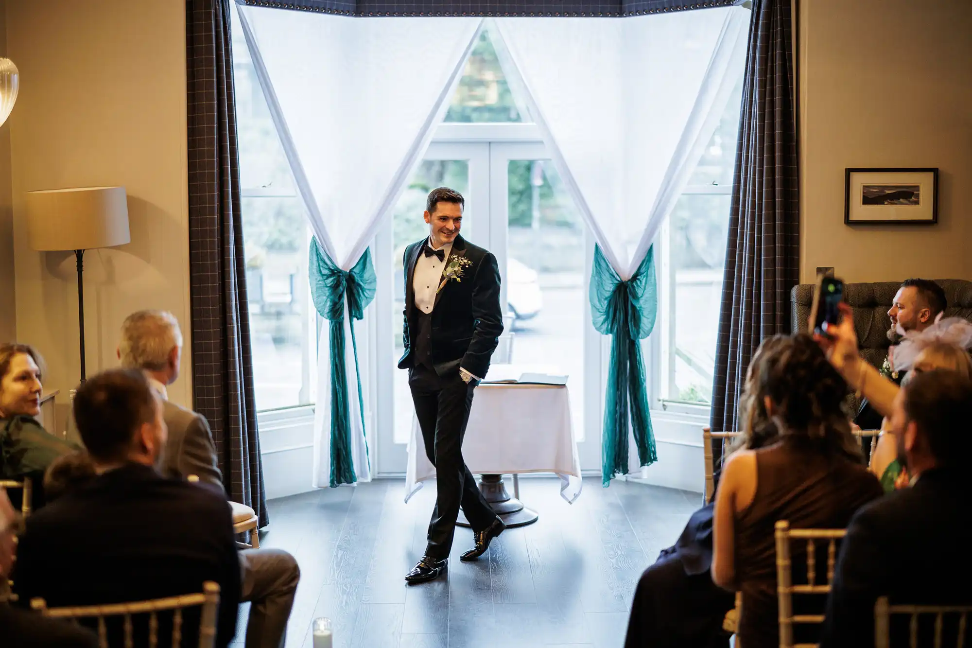 A man in a black tuxedo and bowtie smiles as he walks past draped white curtains with green bows. Guests watch from chairs.