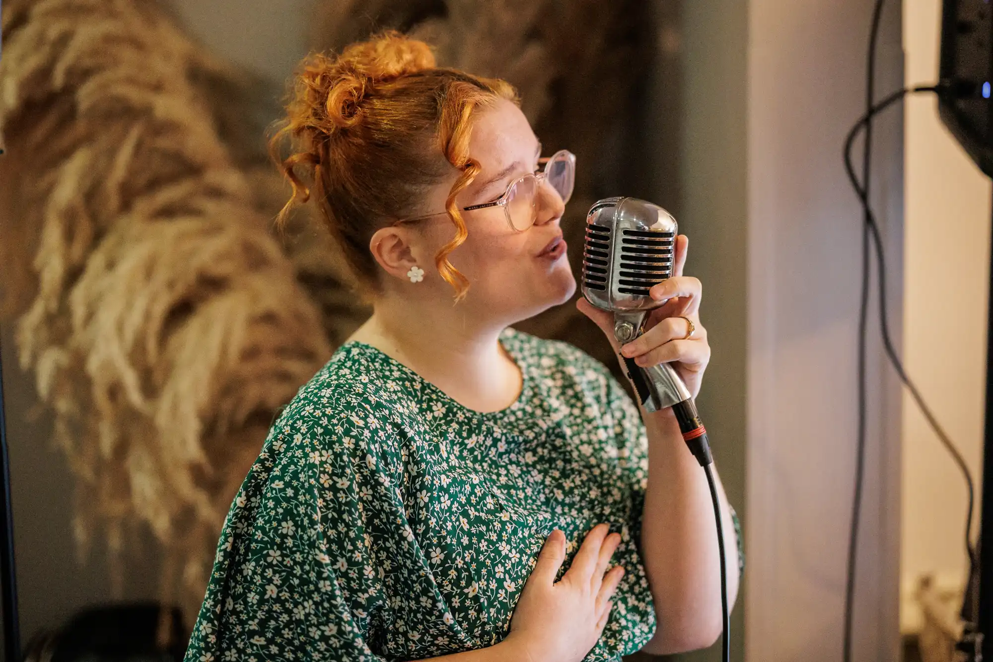 A young woman with red hair pulled up and wearing glasses sings into a vintage microphone. She wears a green floral print dress and has her hand on her chest.