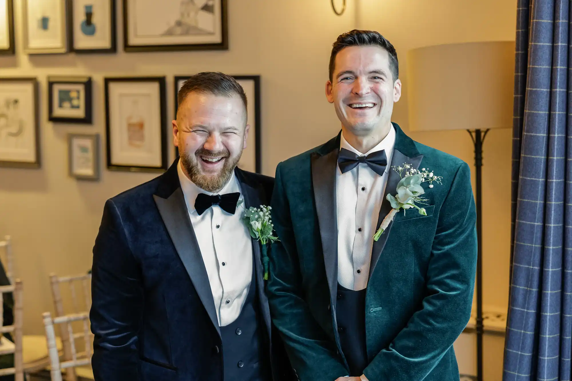 Two grooms in tuxedos laugh joyfully. One wears a navy velvet jacket, the other a teal velvet jacket. Both have boutonnieres and black bow ties.