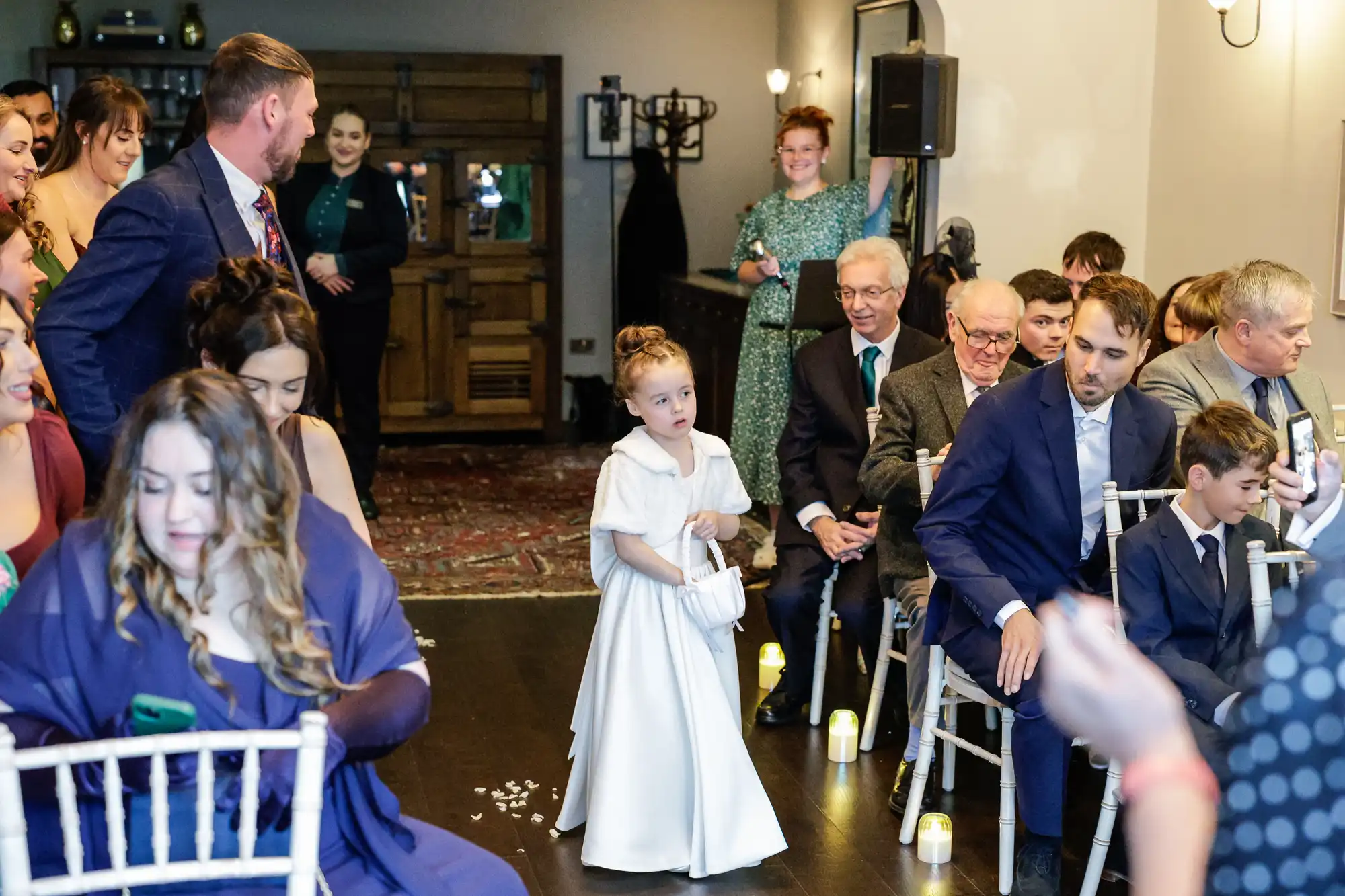 A young girl in a white dress and cape walks down an aisle holding a basket, guests watch as a man in a blue suit sits.
