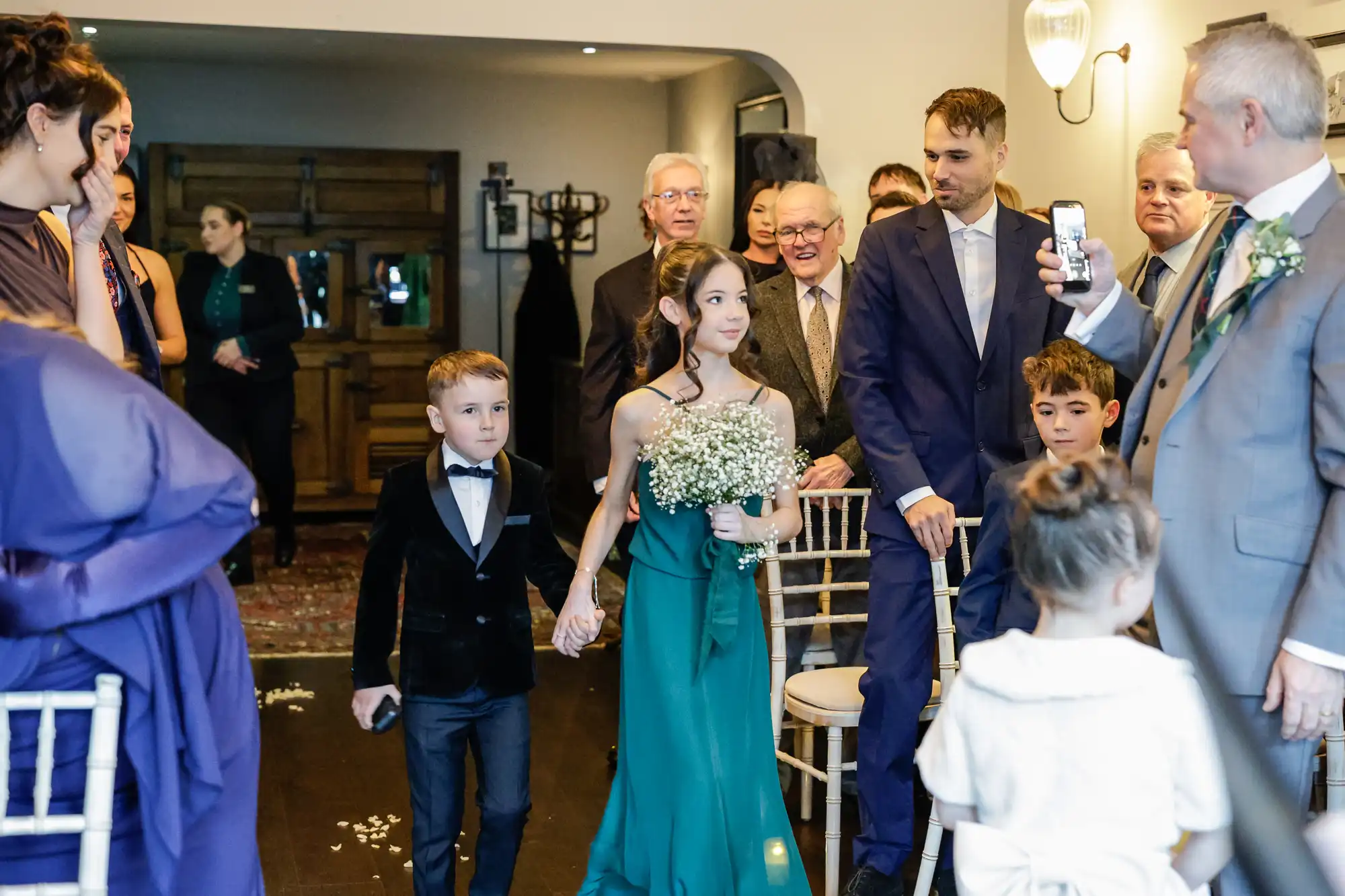 A young boy in a black tuxedo and a girl in a teal dress hold hands walking down an aisle, surrounded by adults in formal wear.