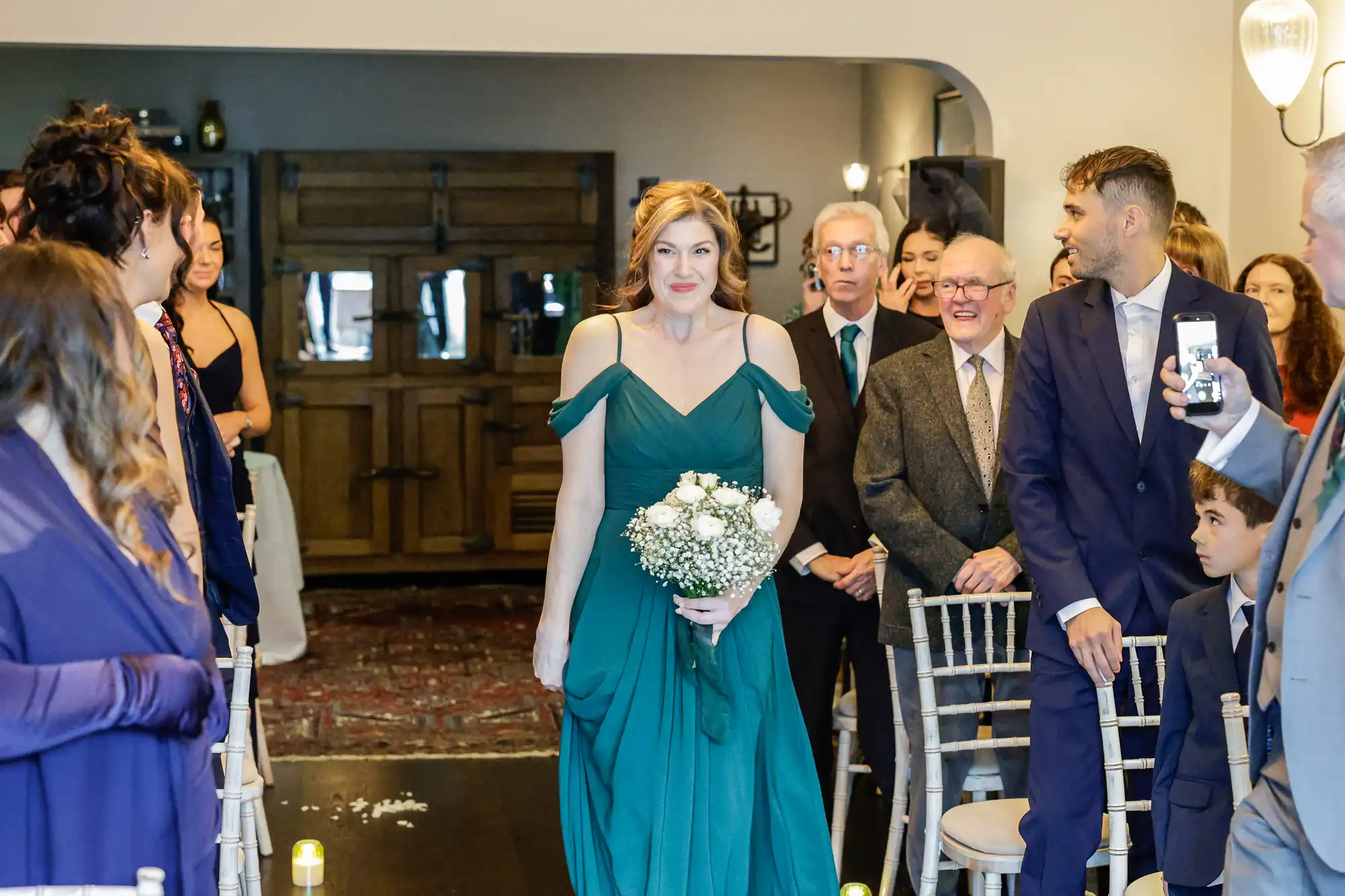A woman in a green off-the-shoulder dress walks down an aisle holding a bouquet of white flowers. She smiles warmly at the camera. Other guests, including men in suits and women in formal attire, are visible in the background.