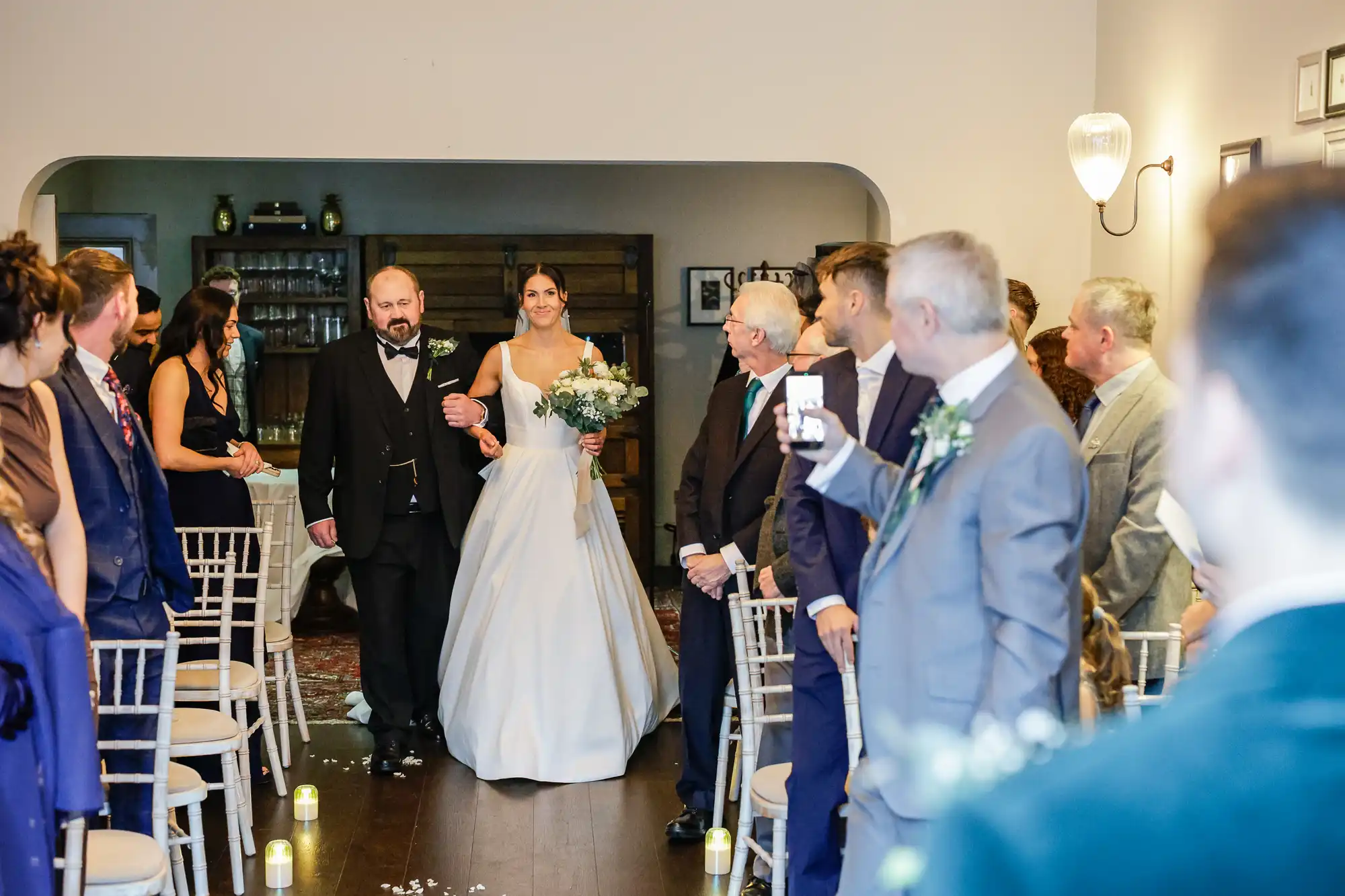 A bride in a white gown walks down an aisle, escorted by a man in a suit. Guests are seated on chairs to the side.