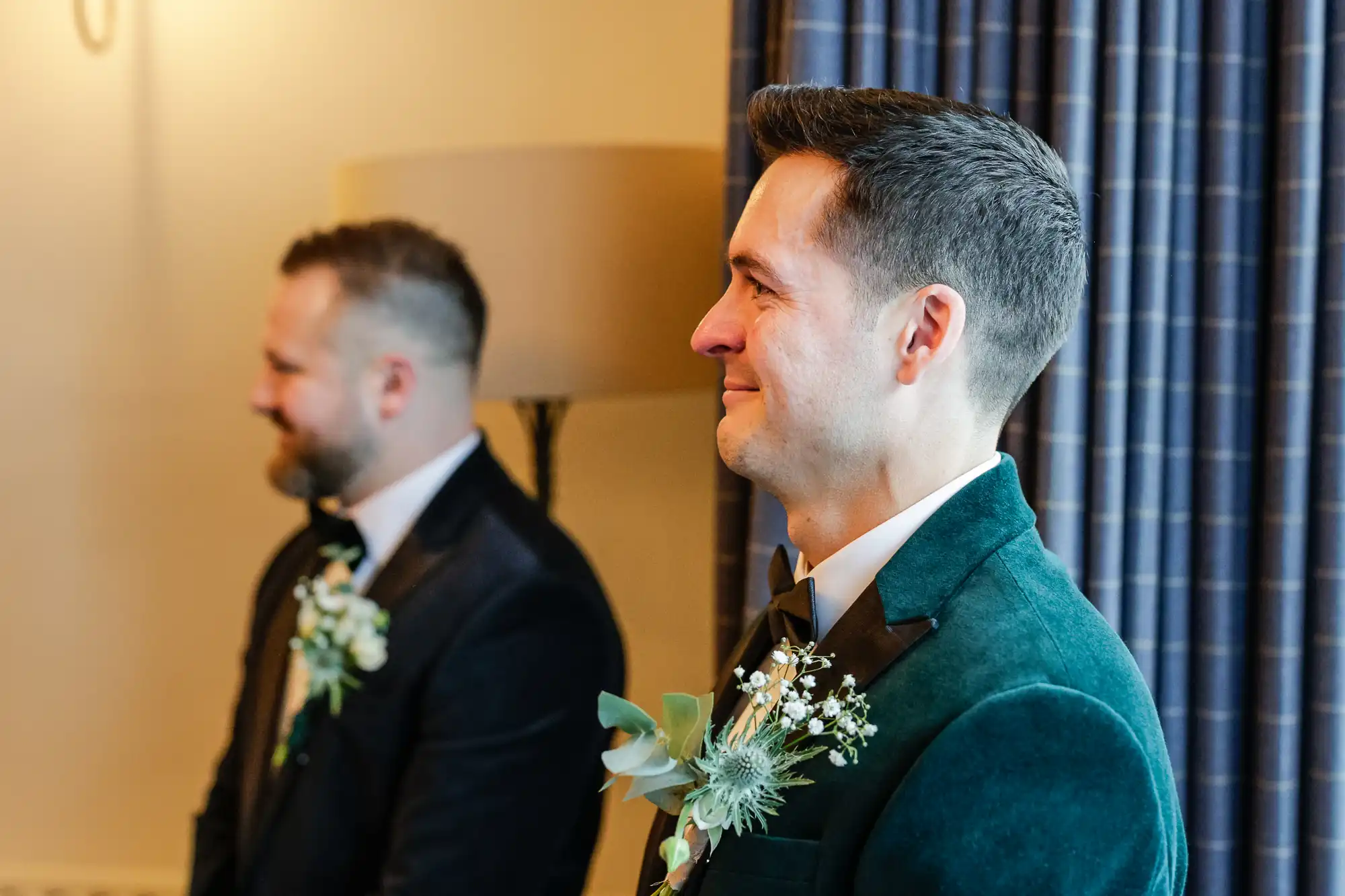 Two men in formal attire with boutonnieres, one in a black suit and the other in a teal velvet jacket, stand with their profiles to the camera.