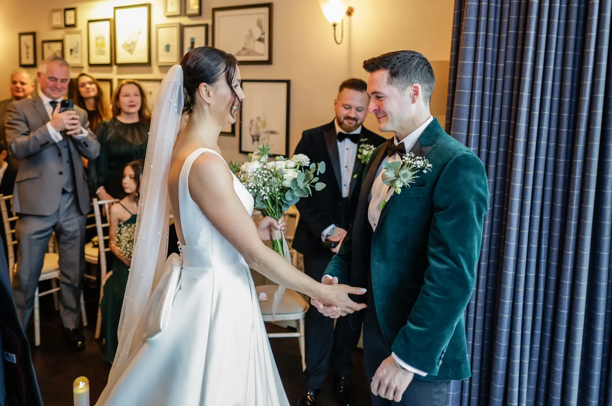 A bride in a white satin dress and veil shakes hands with her groom in a deep green velvet jacket. He smiles back at her as a man in a tuxedo officiates behind them.
