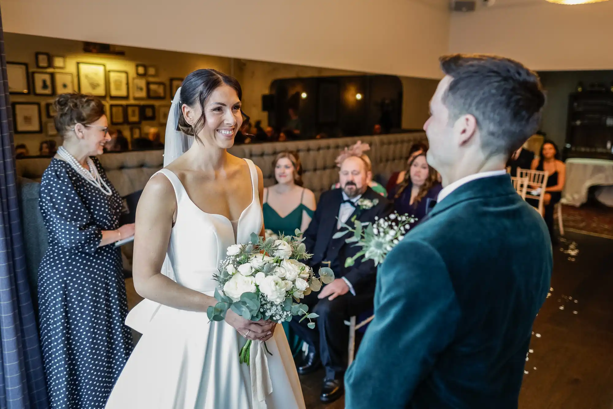 A bride smiles as she holds a bouquet, standing opposite her groom in a teal velvet jacket. Guests and a woman in a polka-dot dress watch.