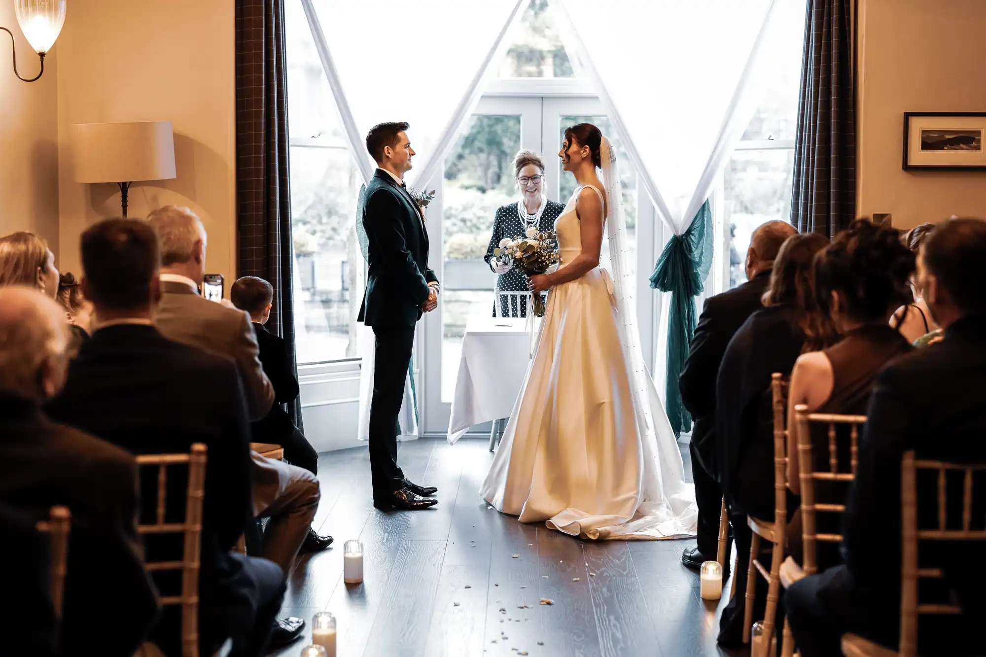 A bride and groom stand facing each other during a wedding ceremony. The bride wears a white gown and veil, holding a bouquet. The groom wears a dark suit. An officiant stands behind them. Guests watch from their seats, with candles on the floor.