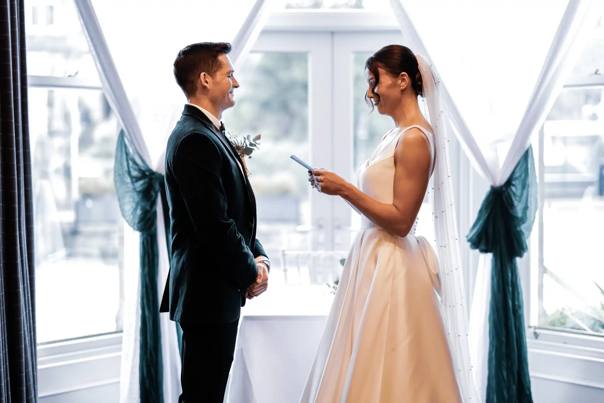 A bride in a white gown reads vows to a groom in a dark suit, both smiling at each other by a window with draped teal fabric.