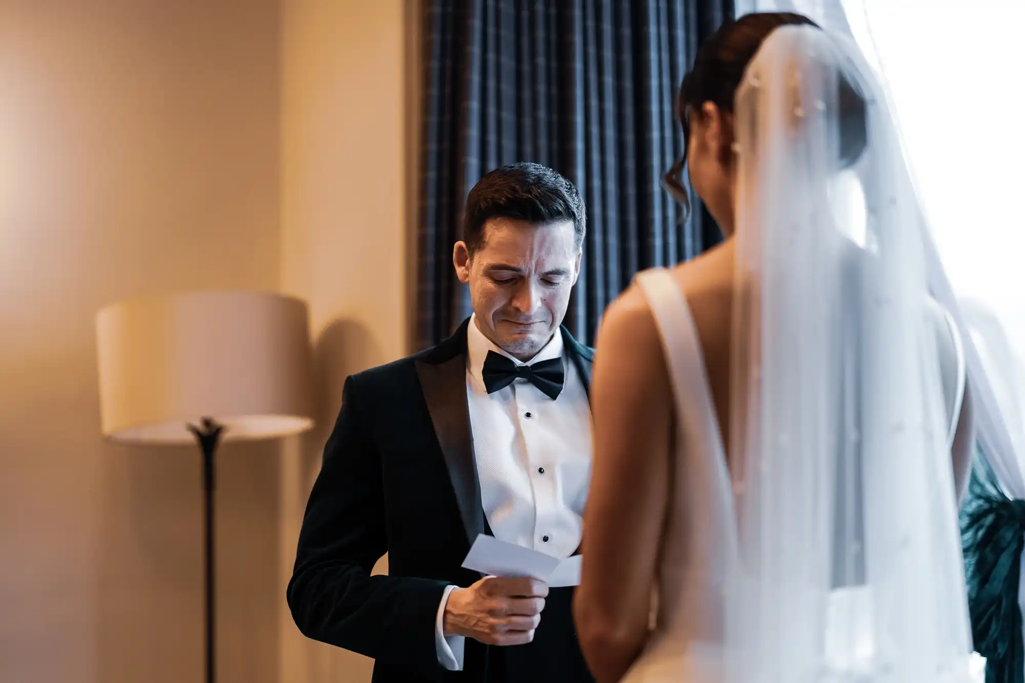 A groom in a tuxedo looks down tearfully at a letter while facing his bride, who is out of focus and wearing a veil and wedding dress.