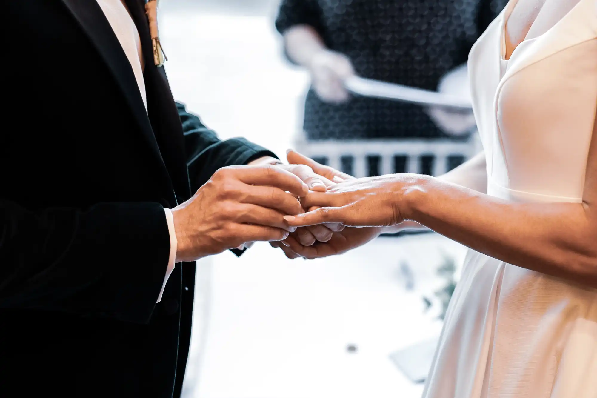 A groom in a black suit places a ring on his bride's finger. The bride wears a white dress. In the background, an officiant holds papers.