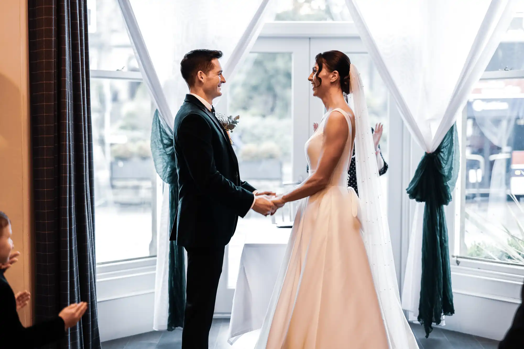 A groom in a dark suit holds hands with a bride in a white wedding dress. They are standing before a large window, with sheer curtains and dark green sashes.
