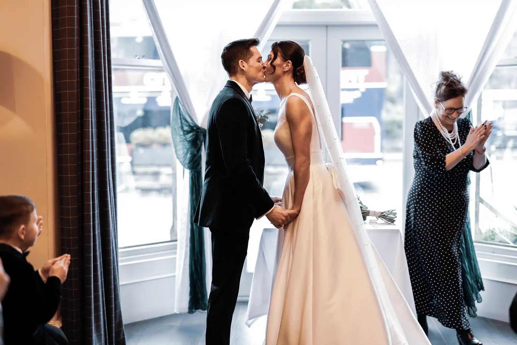 A bride and groom kiss during their wedding ceremony. A woman in a polka dot dress claps her hands in the background. A young boy in a tuxedo also claps.