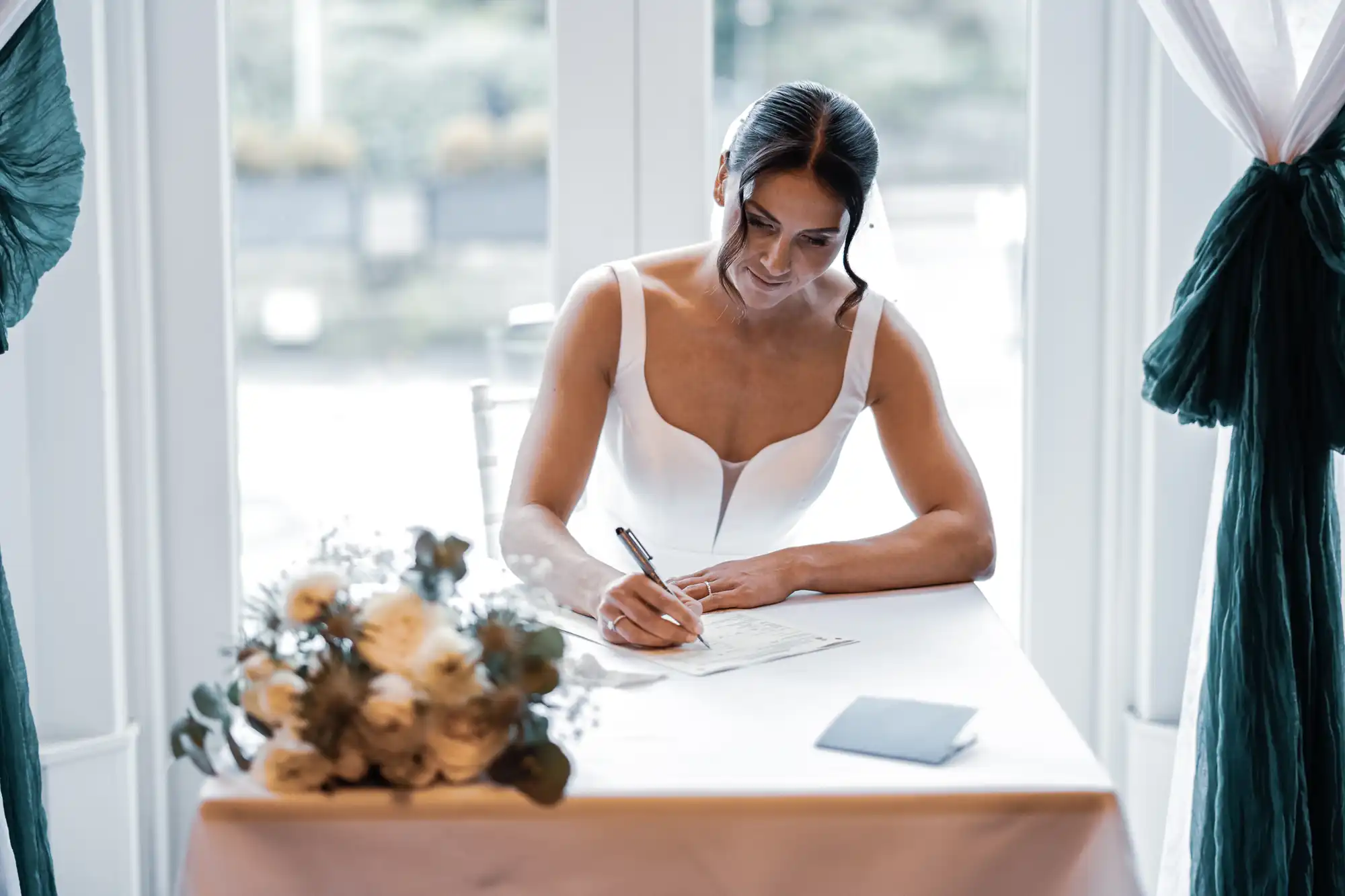 A bride in a white dress signs a document at a table with a bouquet of flowers beside her.