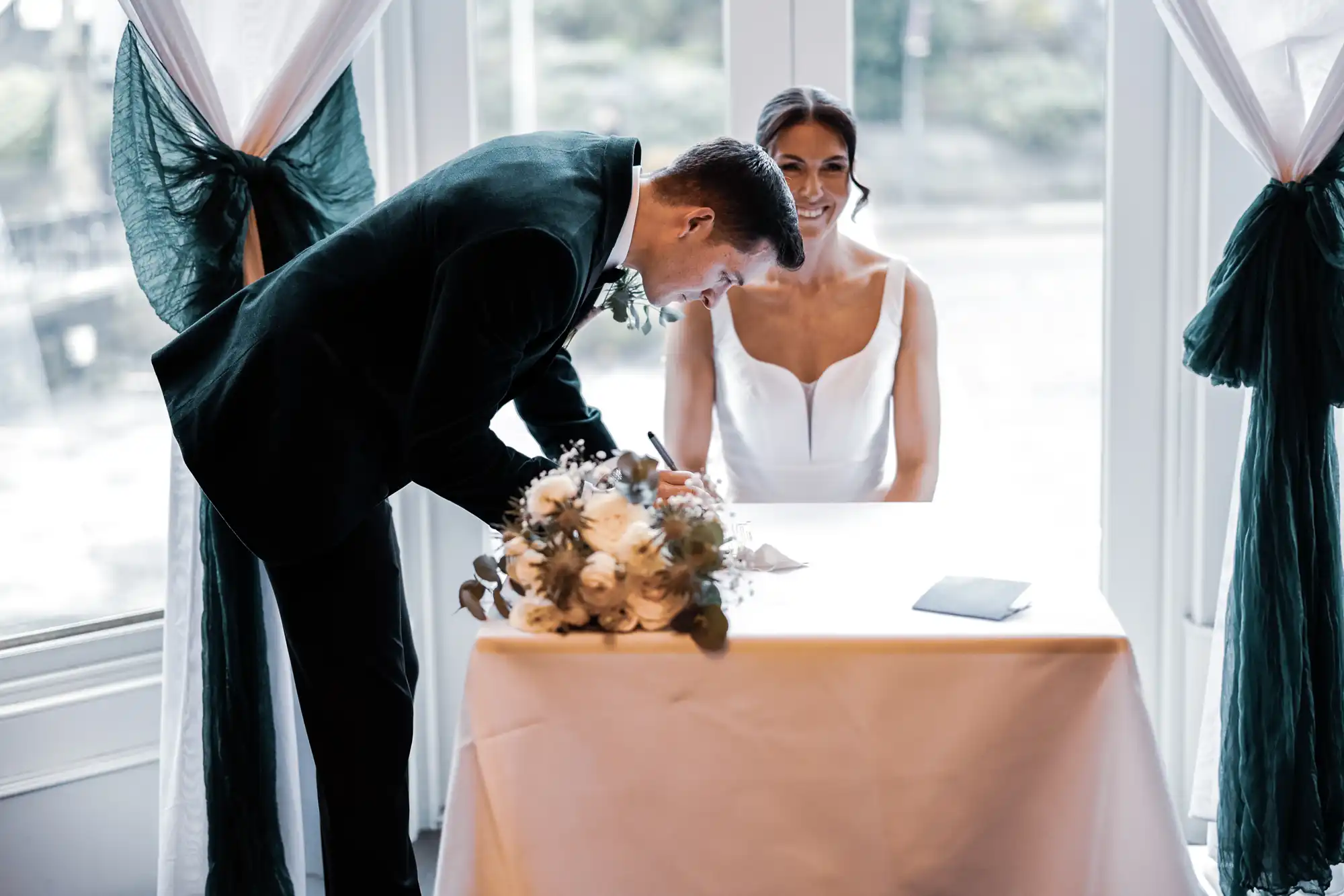 A groom in a dark green suit signs a marriage certificate while his smiling bride in a white dress watches. A bouquet of white flowers rests on the table.