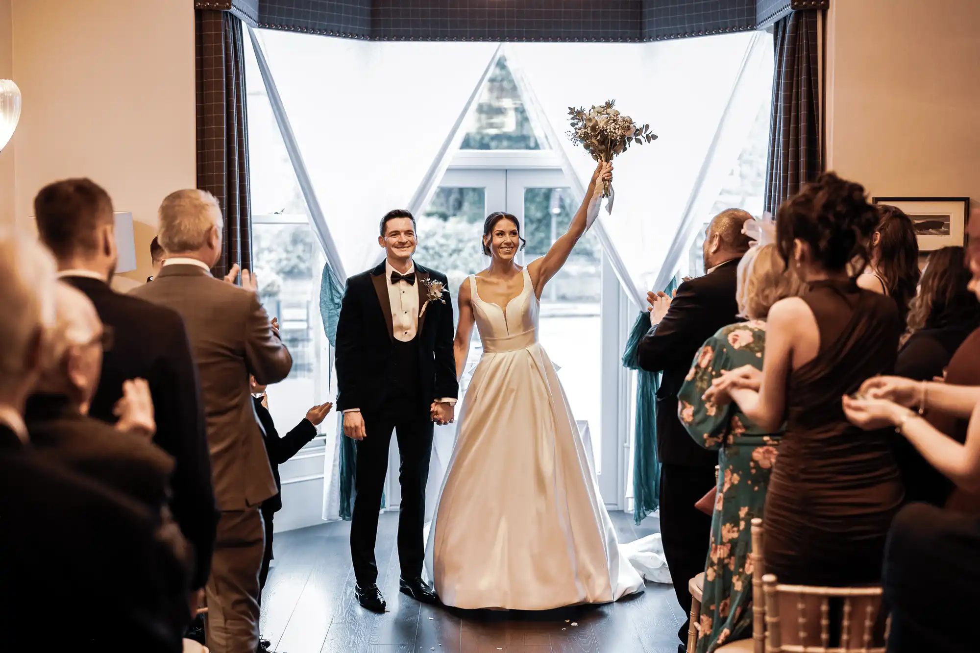 A bride and groom walk down the aisle, holding hands. The bride is in a white wedding gown, holding a bouquet aloft, while the groom is in a tuxedo. Guests are clapping and cheering.