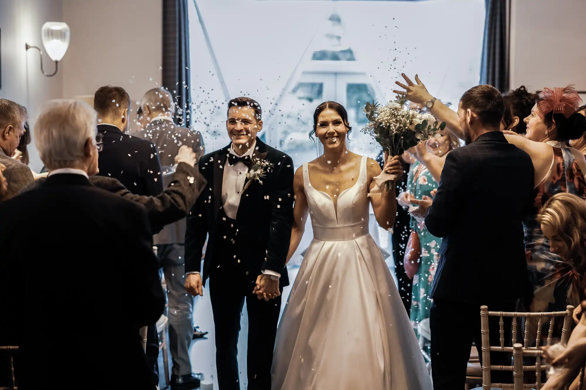 A newlywed couple smiles as they walk through a shower of confetti, surrounded by wedding guests. The bride wears a white satin dress, and the groom a black tuxedo.