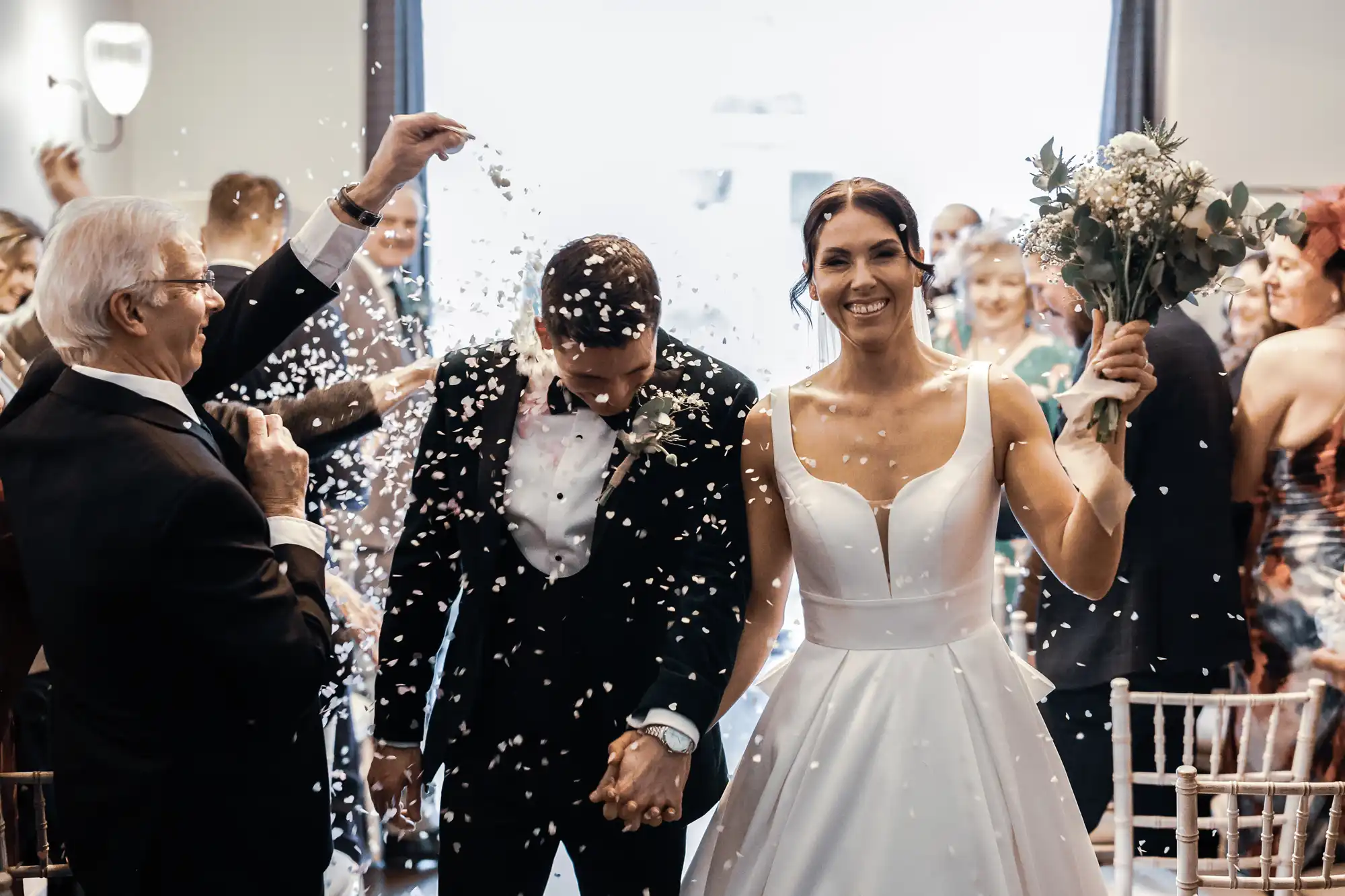 A bride and groom smile as they walk through a shower of confetti, holding hands. The bride wears a white satin dress, and the groom a black tuxedo. Guests in the background celebrate.