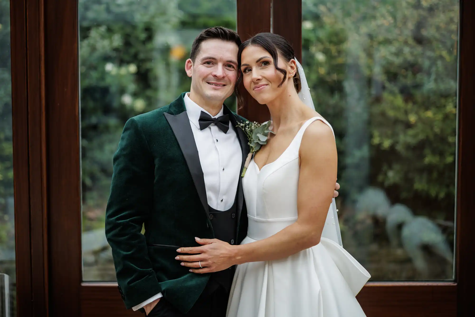 A bride and groom pose together in front of large windows overlooking greenery. The groom wears a dark green velvet tuxedo with a black bow tie; the bride wears a sleeveless white gown and veil, leaning against him smiling.