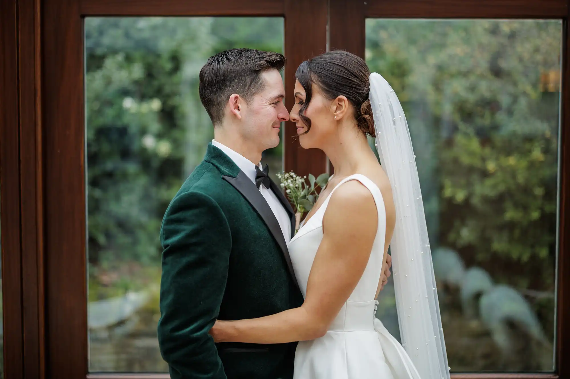 A groom in a green velvet jacket smiles at his bride, who wears a white dress and veil, as they embrace and touch noses in front of a window with a blurred green background.