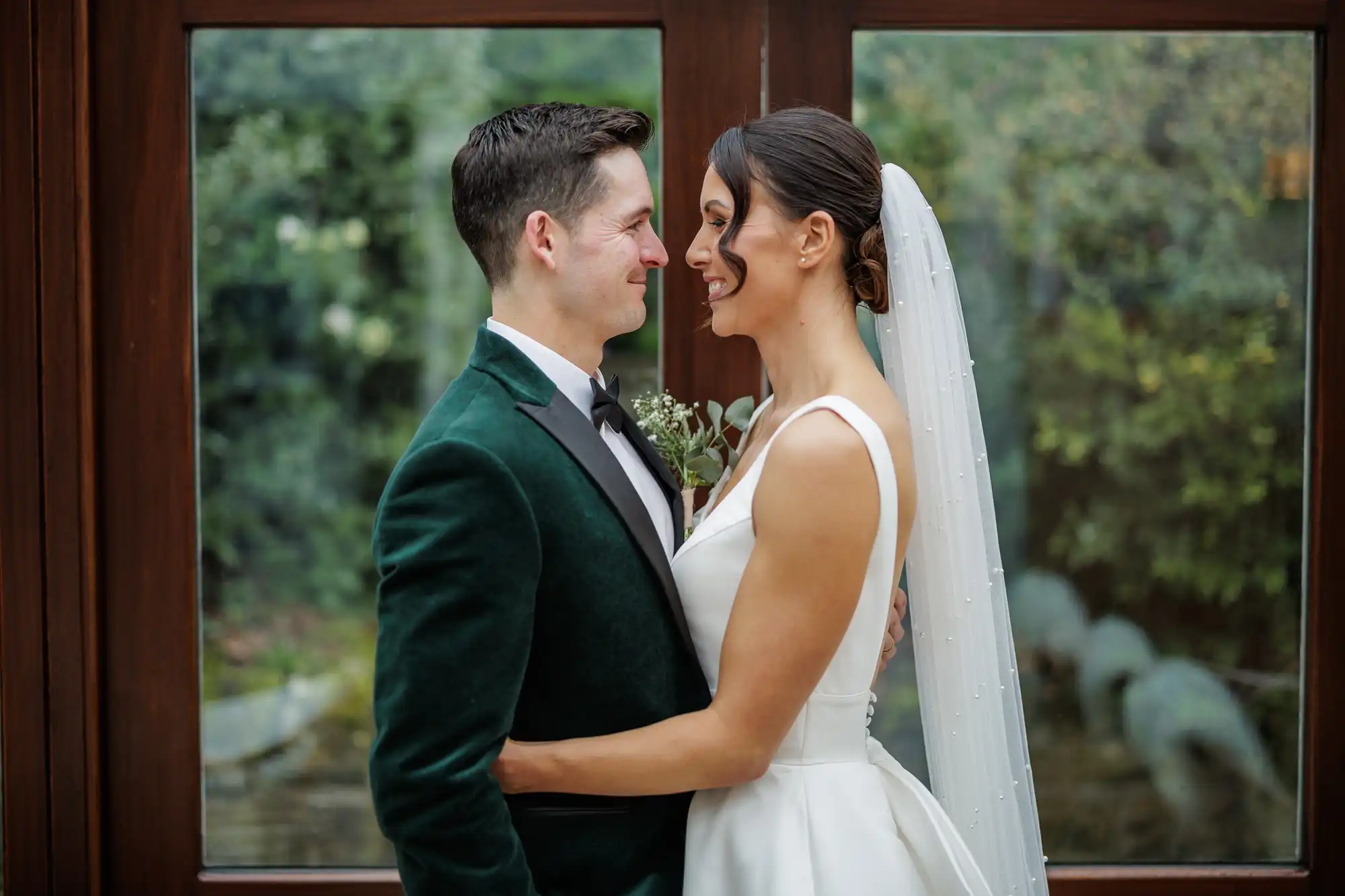 A bride in a white dress and veil smiles at her groom, who wears a green velvet suit. They embrace by a window overlooking greenery.