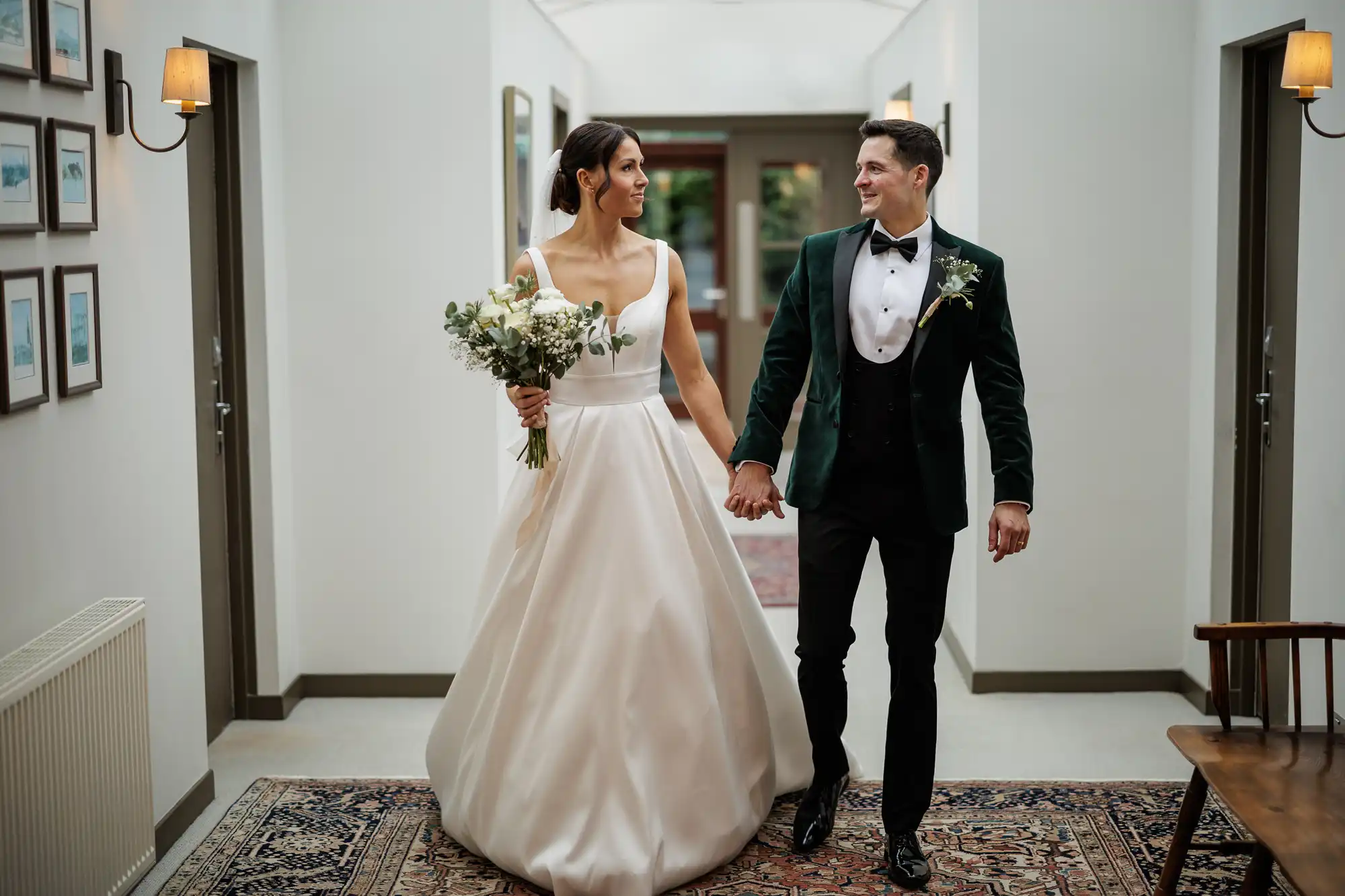 A bride and groom walk hand-in-hand down a hall. She wears a satin gown and carries a bouquet. He wears a dark green velvet tuxedo.