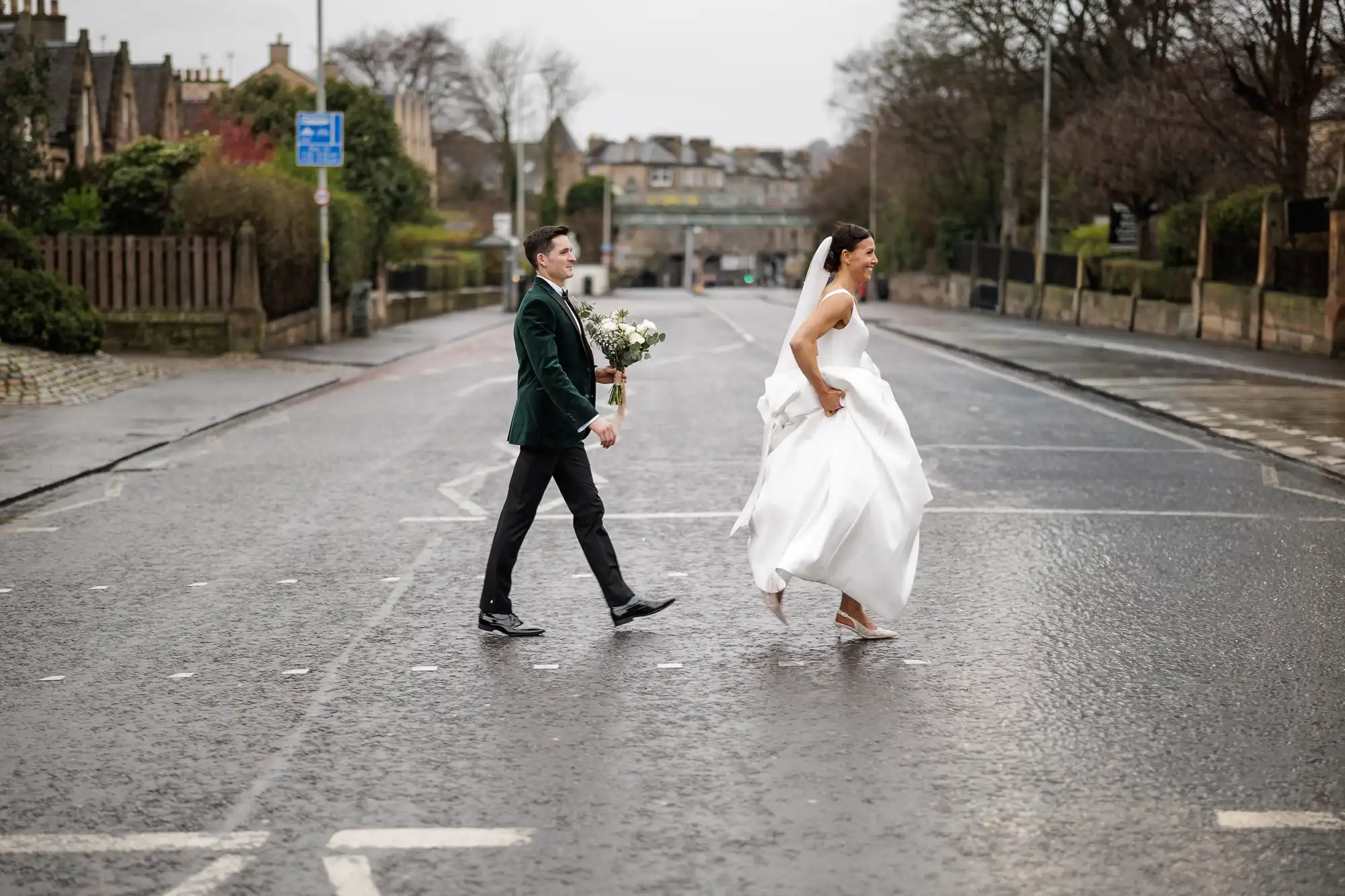 A groom in a green jacket and a bride in a white dress walk across a wet street, holding hands and smiling.