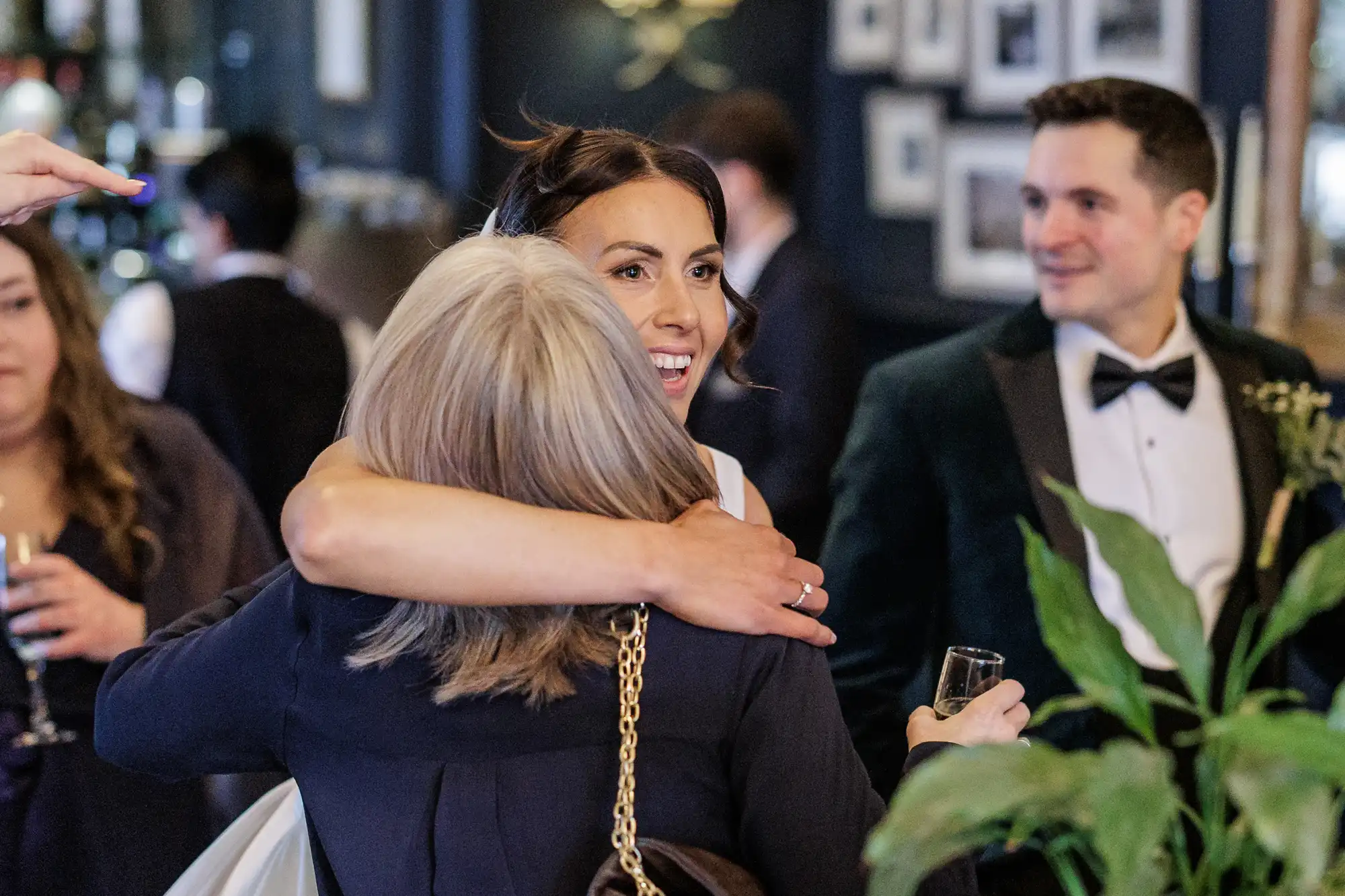 A bride in a white dress hugs a woman from behind. The bride laughs joyfully, her eyes sparkling. Behind them, a groom in a dark green suit smiles.