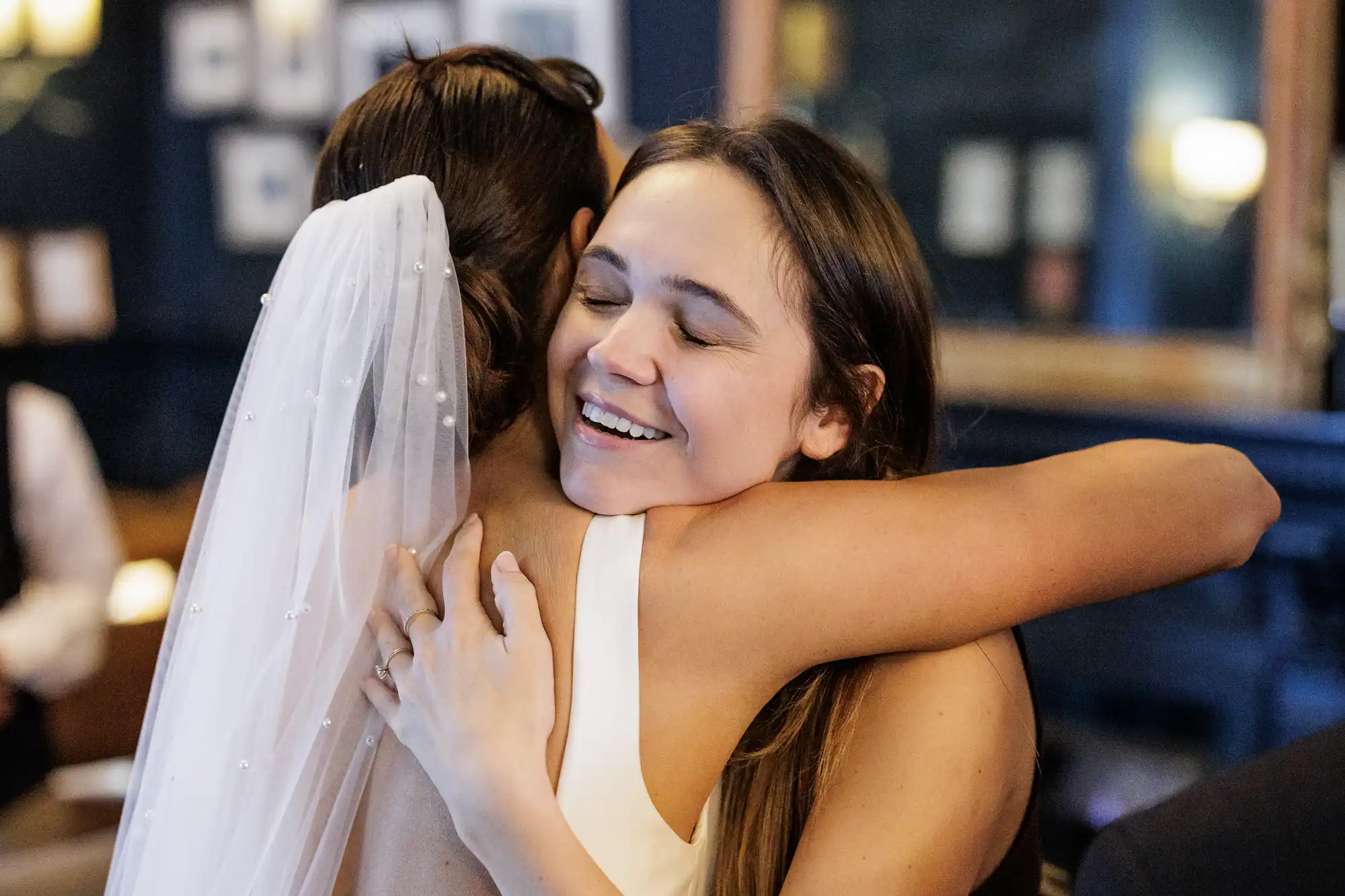 A bride in a pearl-adorned veil hugs a smiling woman wearing a white dress. The woman's eyes are closed as she embraces the bride tightly, her arms wrapped around the bride's shoulders.