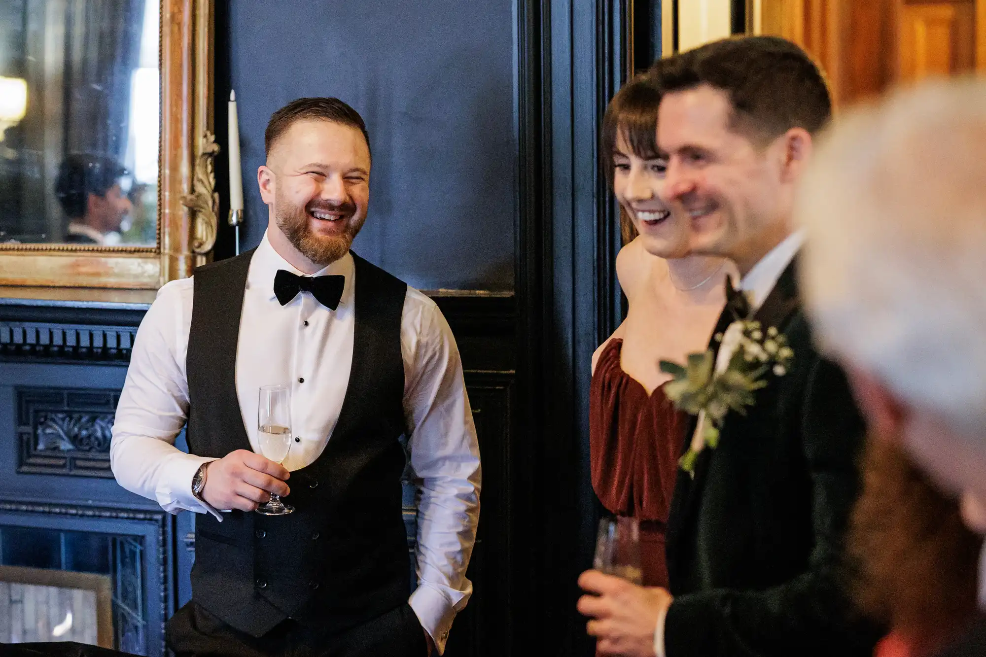 A groom in a tuxedo smiles while holding a champagne flute, with a bride in a red dress and another man in a dark suit behind him at a wedding reception.