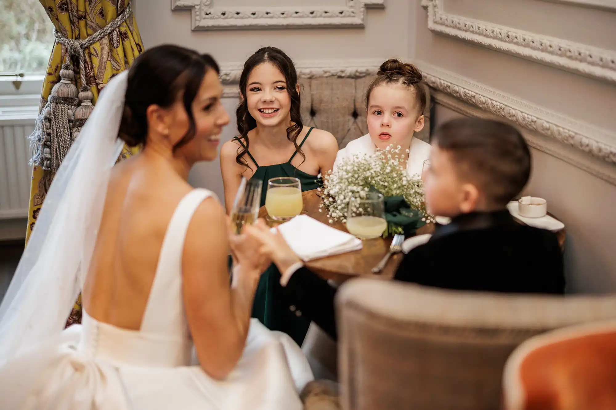 A bride in a white dress raises a champagne flute, smiling. She sits with two girls in green dresses and a boy in a black suit at a table with drinks and a bouquet of baby's breath.