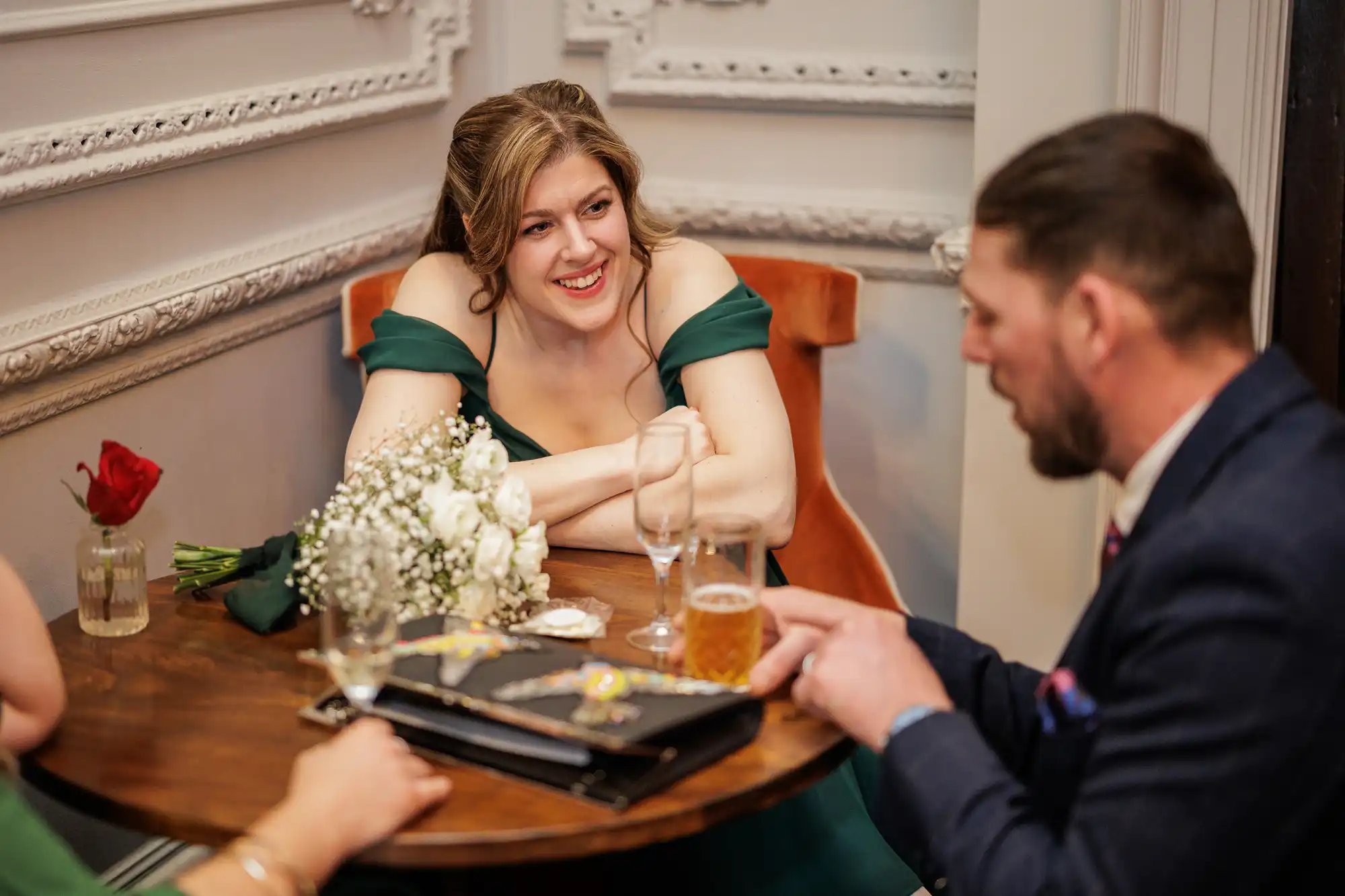 A woman in a green dress smiles while sitting at a round wooden table with a man in a suit. A bouquet of white flowers and a single red rose in a vase are also on the table.