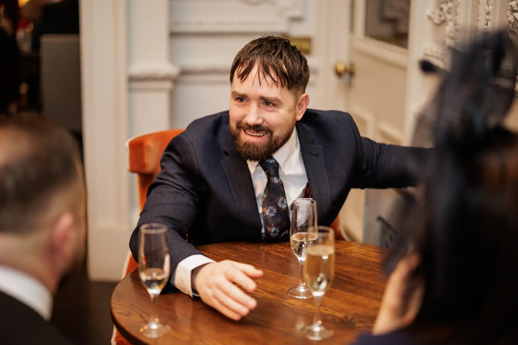 A smiling man in a dark suit and floral tie sits at a round wooden table with two glasses of champagne.