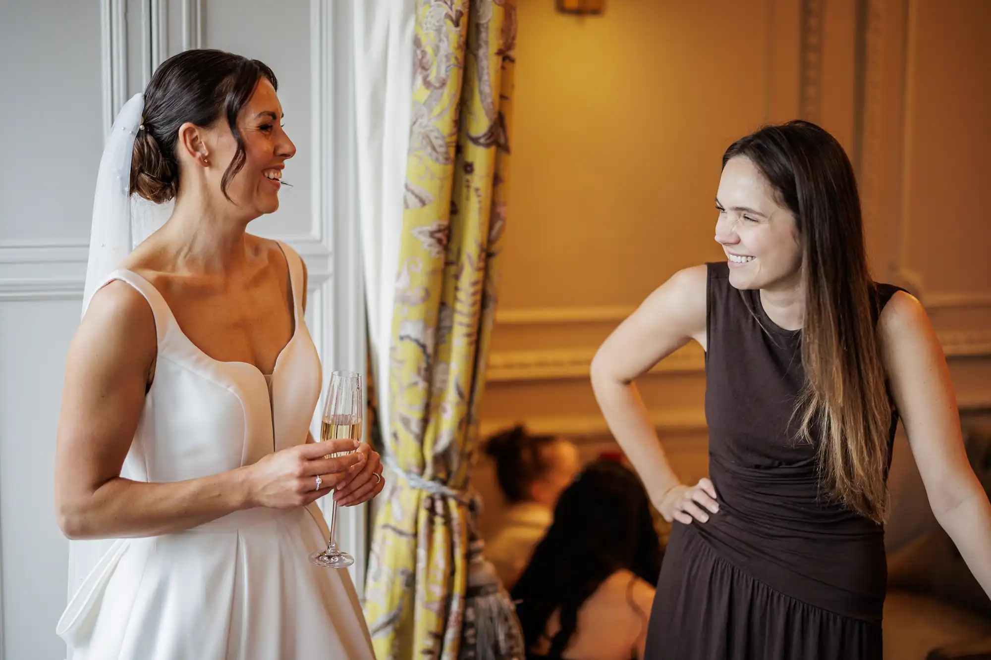 A bride in a white gown and veil smiles at a woman in a dark brown dress, both holding champagne glasses.