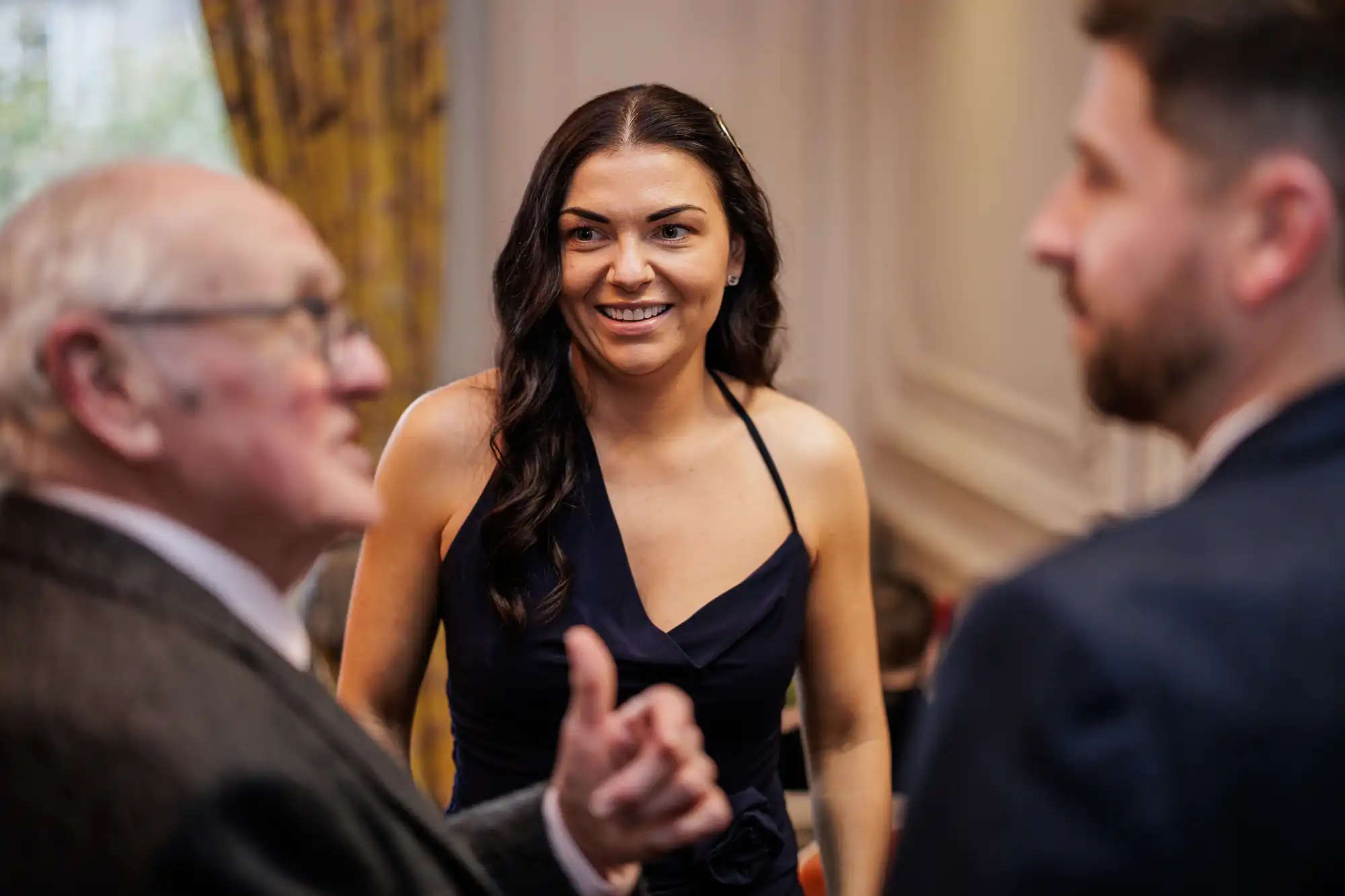 A woman in a dark blue halter dress smiles between two men. One man, on the left, is older, with glasses, and is in profile facing away. The other man, on the right, is in profile facing away.