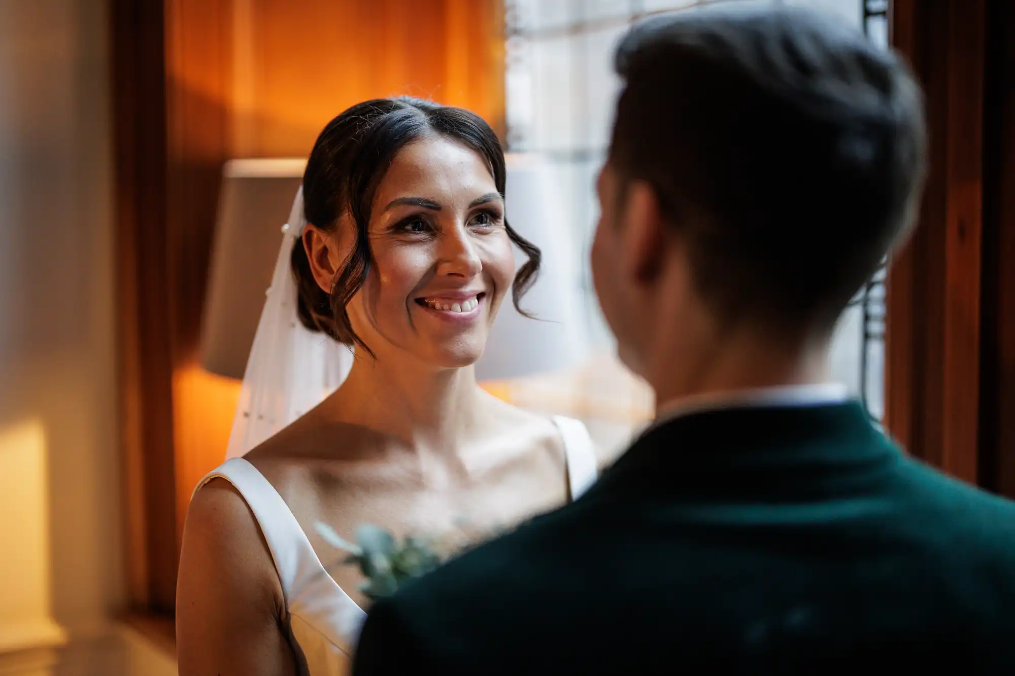 A bride in a white dress and veil smiles warmly at her groom, whose back is to the camera.