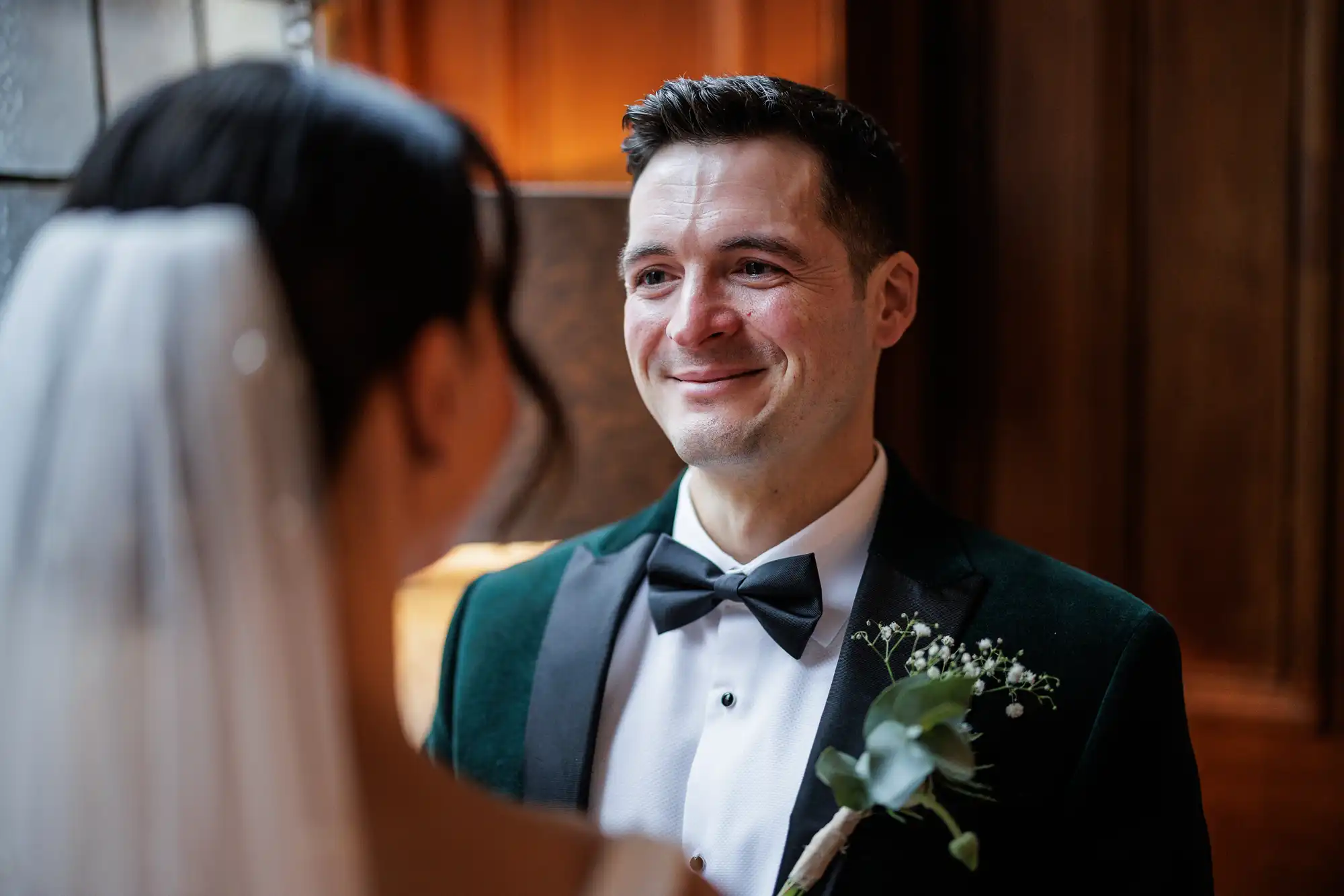 A groom in a green velvet tuxedo smiles lovingly at his bride, who is mostly out of frame with her veil visible.