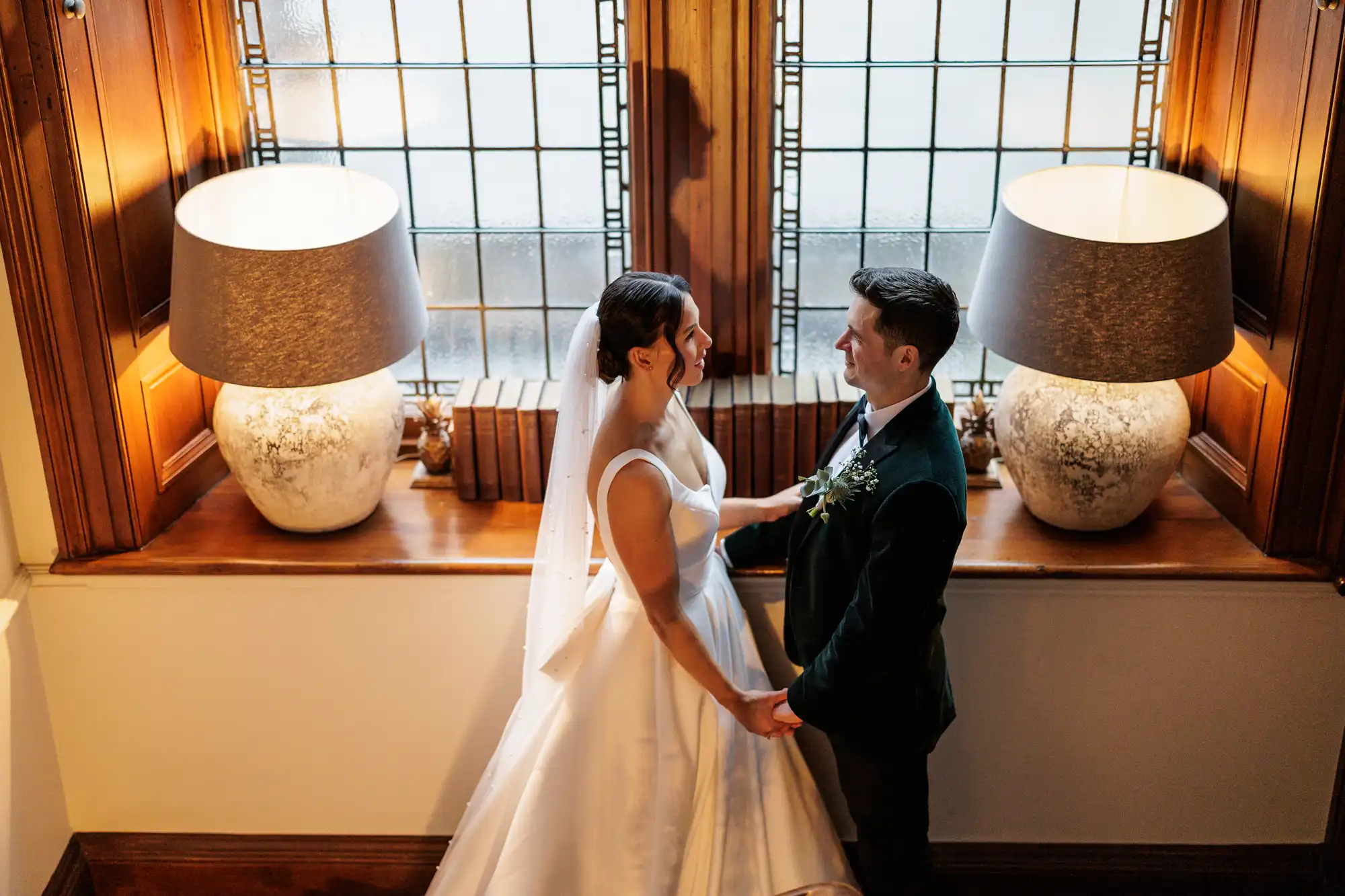 A bride in a white gown and veil holds hands with a groom in a dark green suit, looking at each other near a window with two lamps.