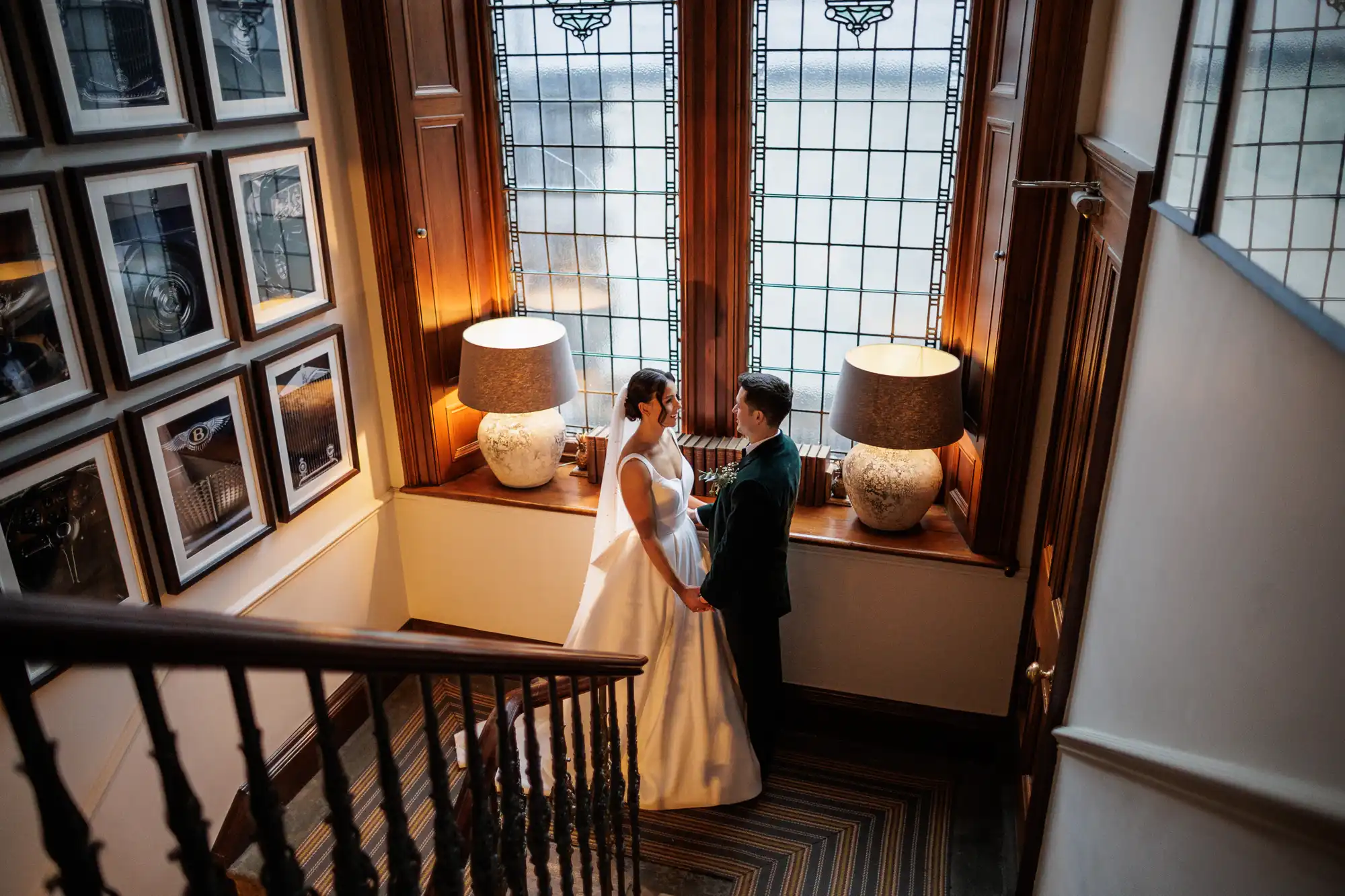 A bride in a white gown and a groom in a dark suit hold hands in front of a large, leaded glass window. They stand between two ornate lamps on a wooden sill.